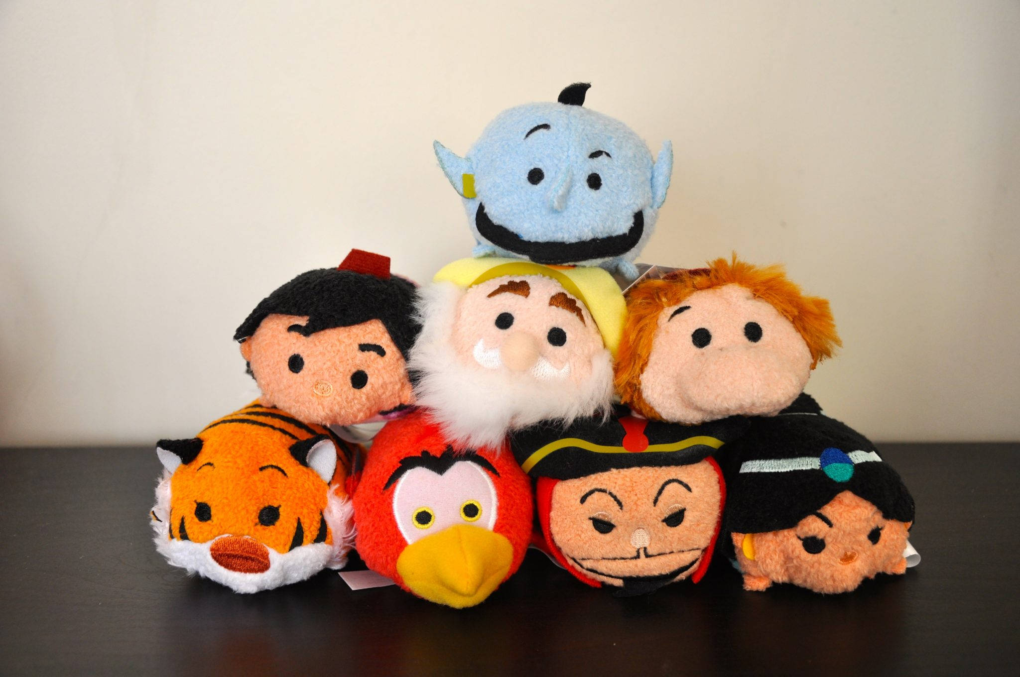 Tsum Tsum 阿拉丁 壁紙 — 一堆色彩繽紛的迪士尼 Tsum Tsum 毛絨玩具，包括阿拉丁、精靈和阿布等角色，排列在深色木質表面上。