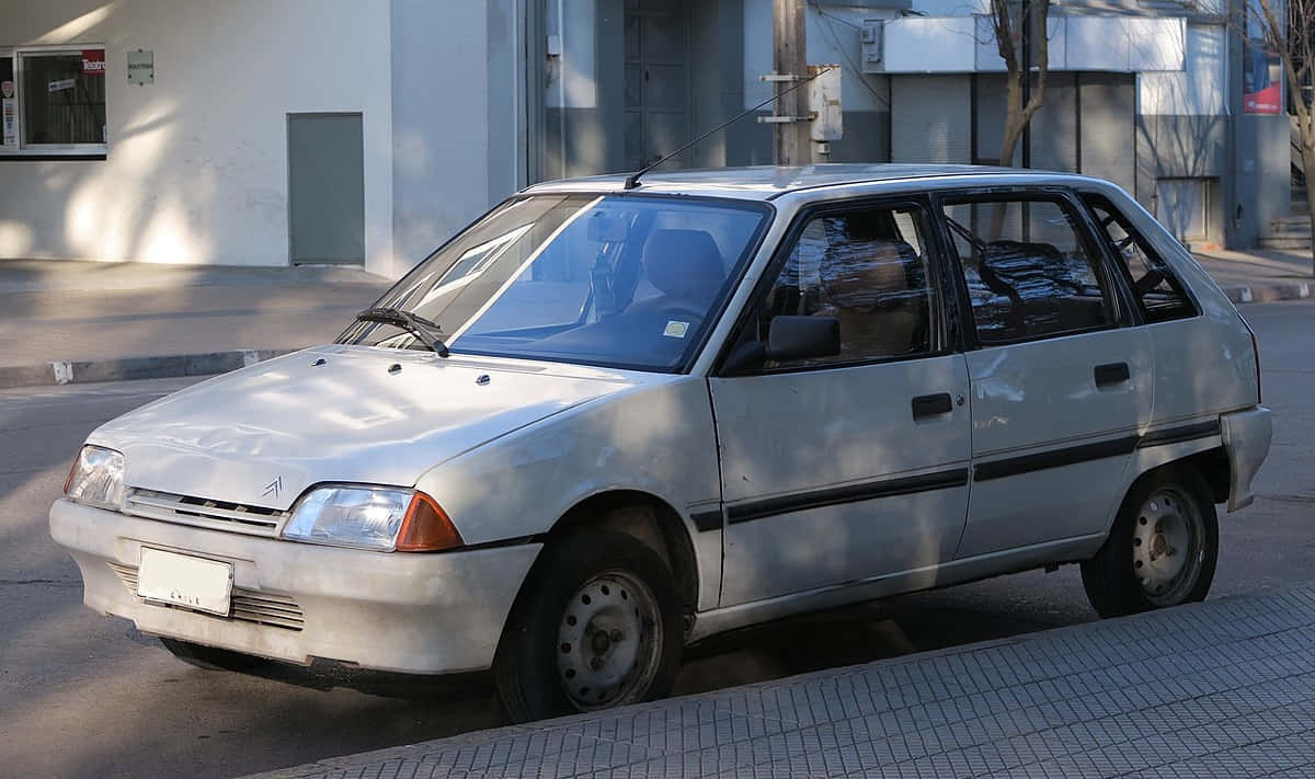  壁紙 — 一輛褪色的銀色掀背車停泊在陽光普照的城市行人路上，旁邊是一座現代建築。