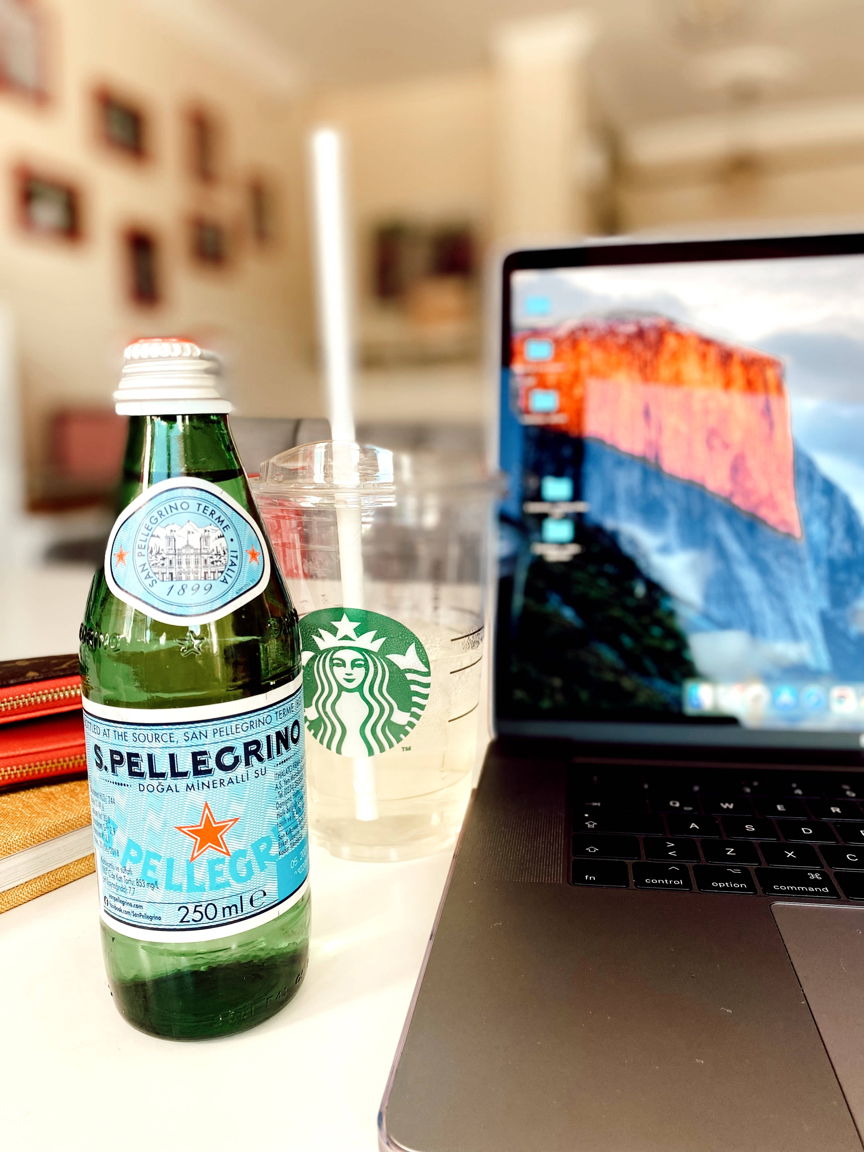 Tumblr風格筆電旁氣泡水 壁紙 — 一個綠色S Pellegrino瓶子和一個Starbucks杯放在一張白色桌子上，旁邊是一部打開的筆記型電腦，顯示著山脈壁紙。