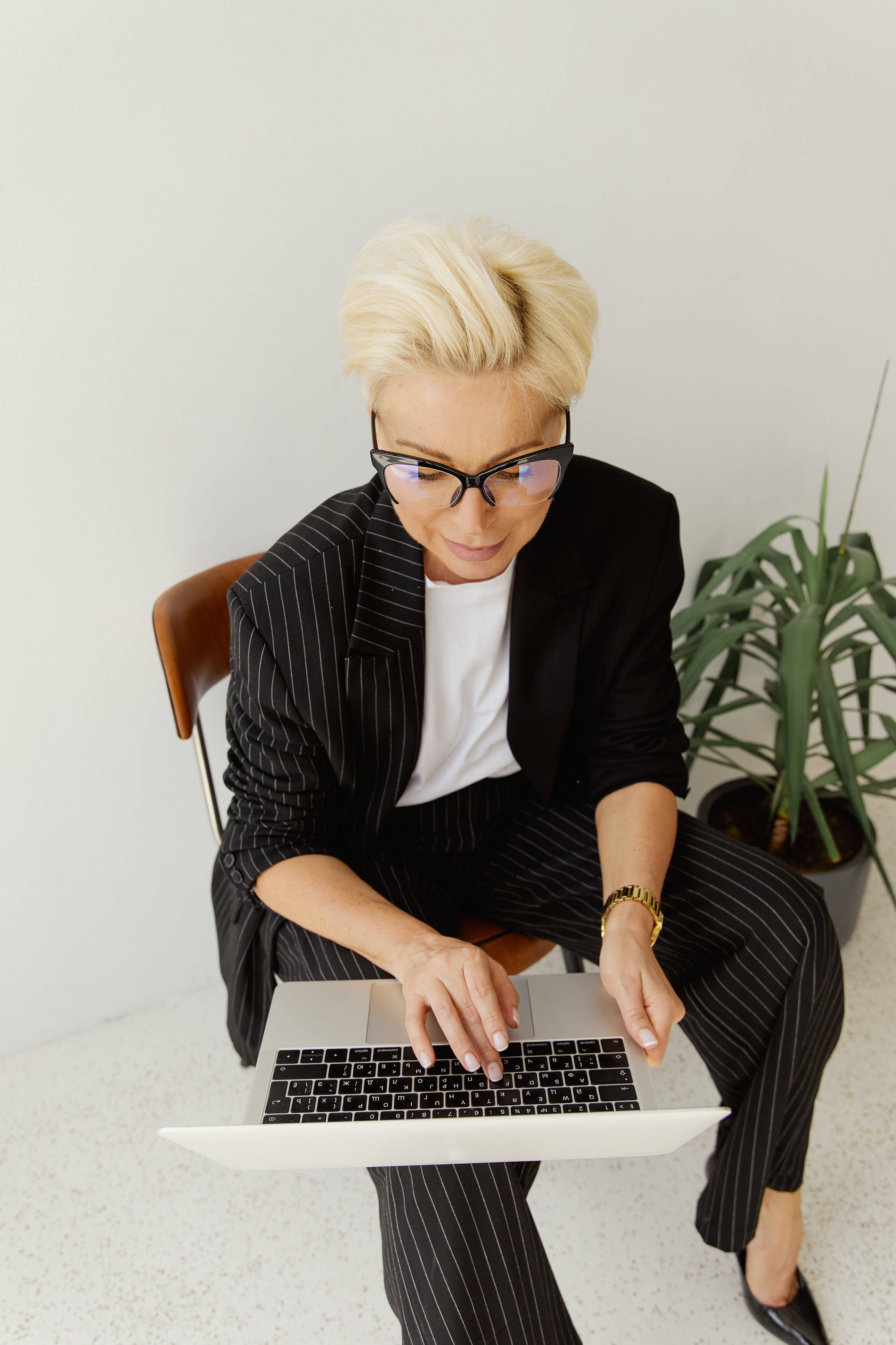 美學Tumblr筆電企業人士 壁紙 — 一位短金髮戴眼鏡的女性坐在簡約風格的房間裡，旁邊有一盆盆栽，正在使用手提電腦打字。