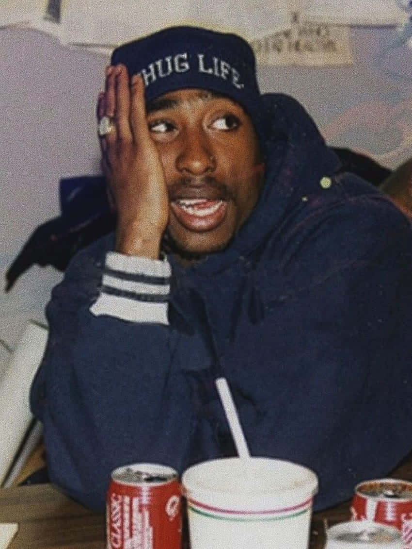  壁紙 — Tupac Shakur 戴著 'HUG LIFE' 帽子和藍色連帽衫，坐在一張有汽水杯的桌子旁，手靠近臉部向上看。