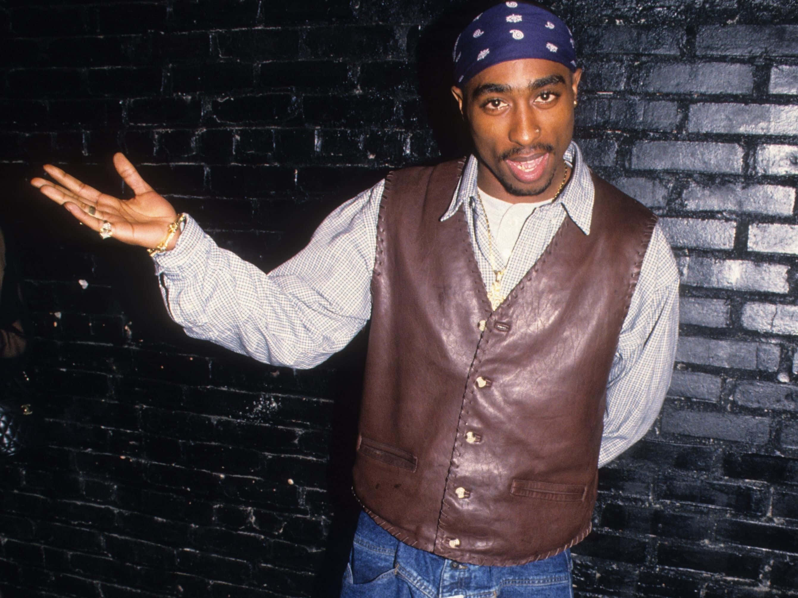 Tupac 桌布 壁紙 — Tupac Shakur 自信地擺姿勢，背景係黑色磚牆，身穿棕色皮背心同藍色頭巾。