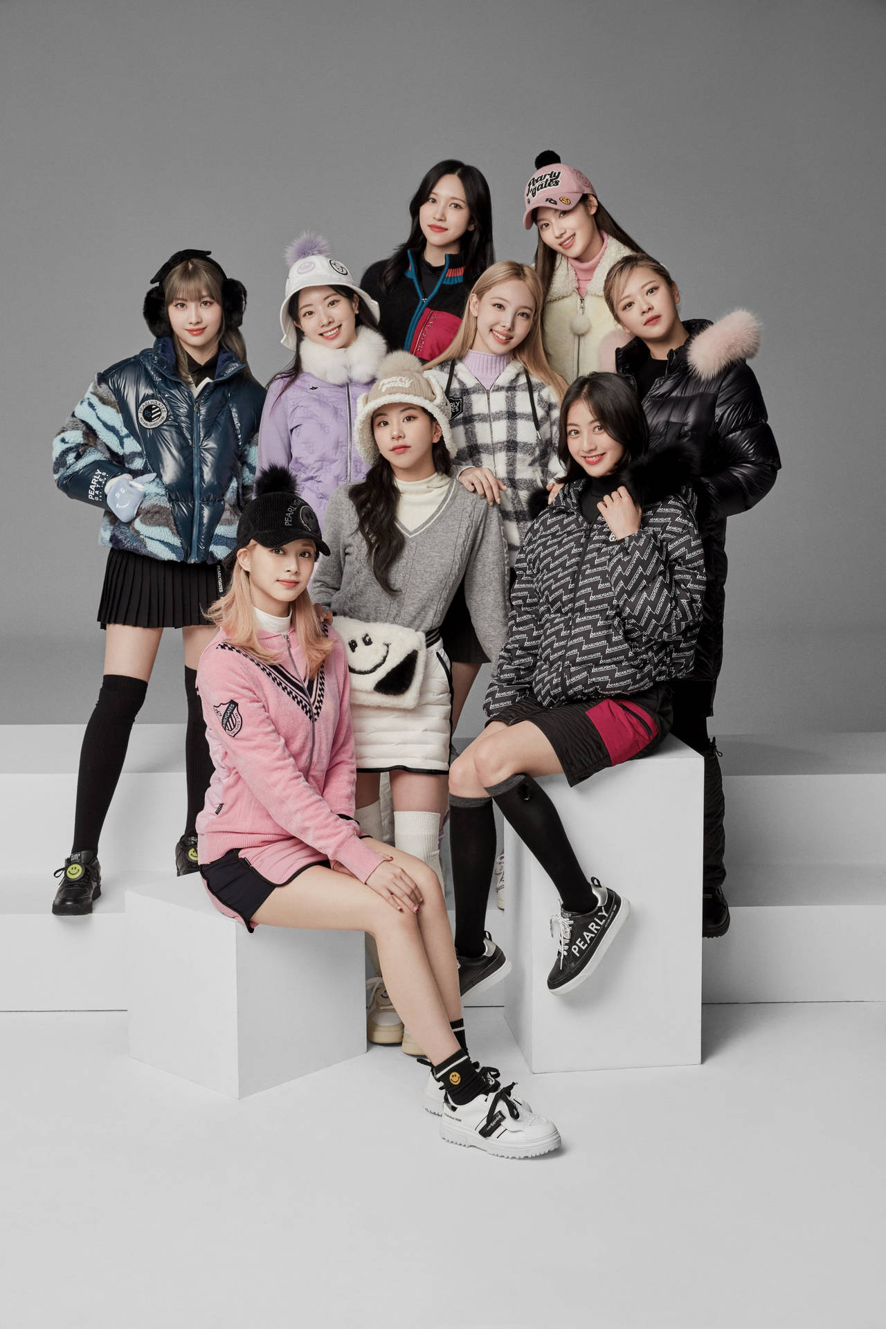 TWICE 4K 冬季服裝 壁紙 — 九位時尚年輕女性穿著潮流冬季外套和配飾，在白色方塊上自信擺姿勢，背景為中性灰色攝影棚。