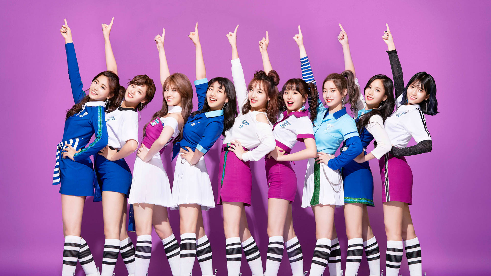 Twice 4K 指向上方姿勢 壁紙 — 九位微笑的女性偶像穿著協調的藍色和粉紅色服裝，在純紫色背景前舉起手臂擺姿勢。