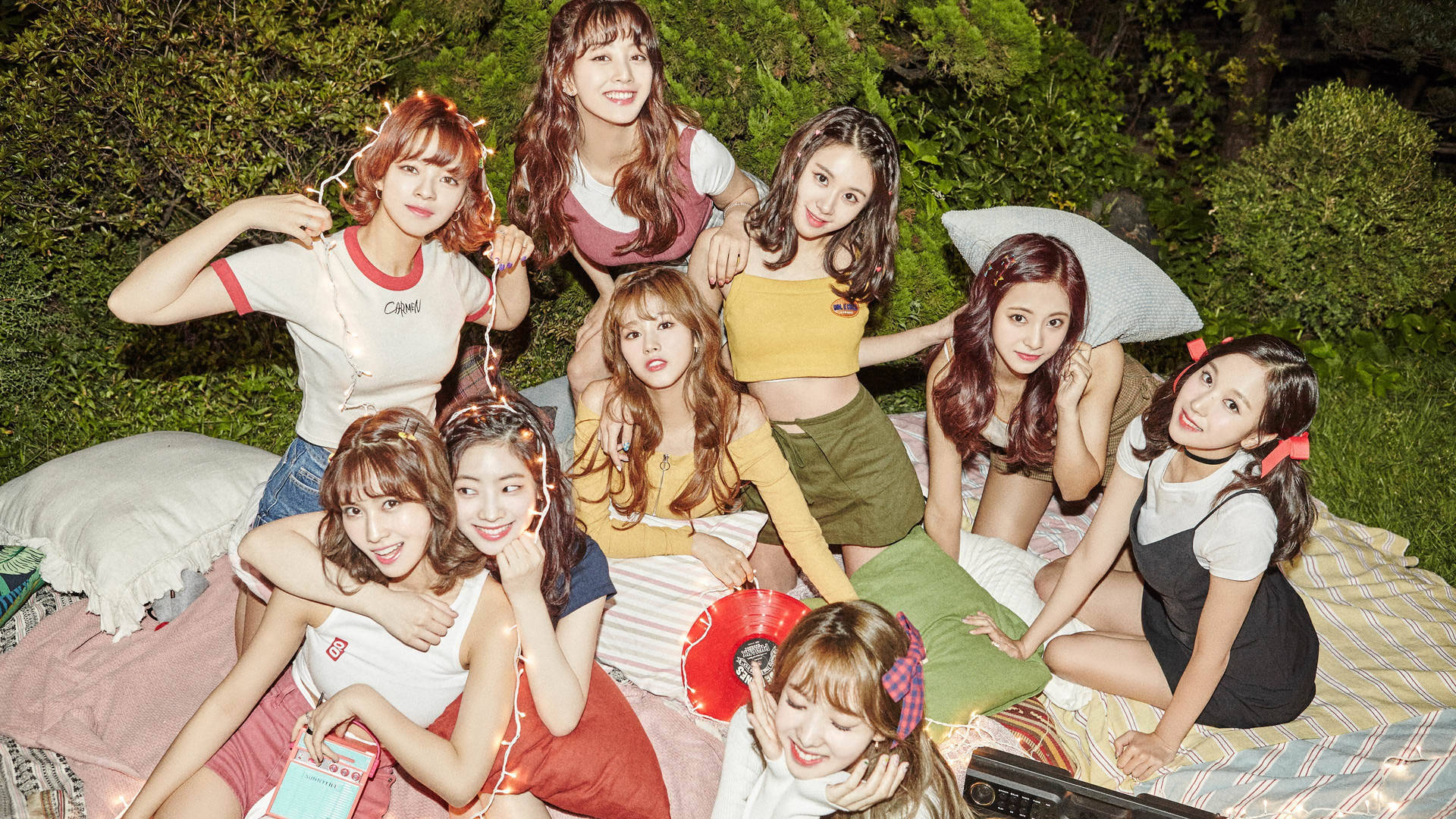 Twice 4K 枕頭戰 壁紙 — 七位微笑的年輕女性穿著色彩繽紛的休閒服裝，在夜間戶外一塊有圖案的毯子上一起擺姿勢，周圍環繞著綠植和燈串。