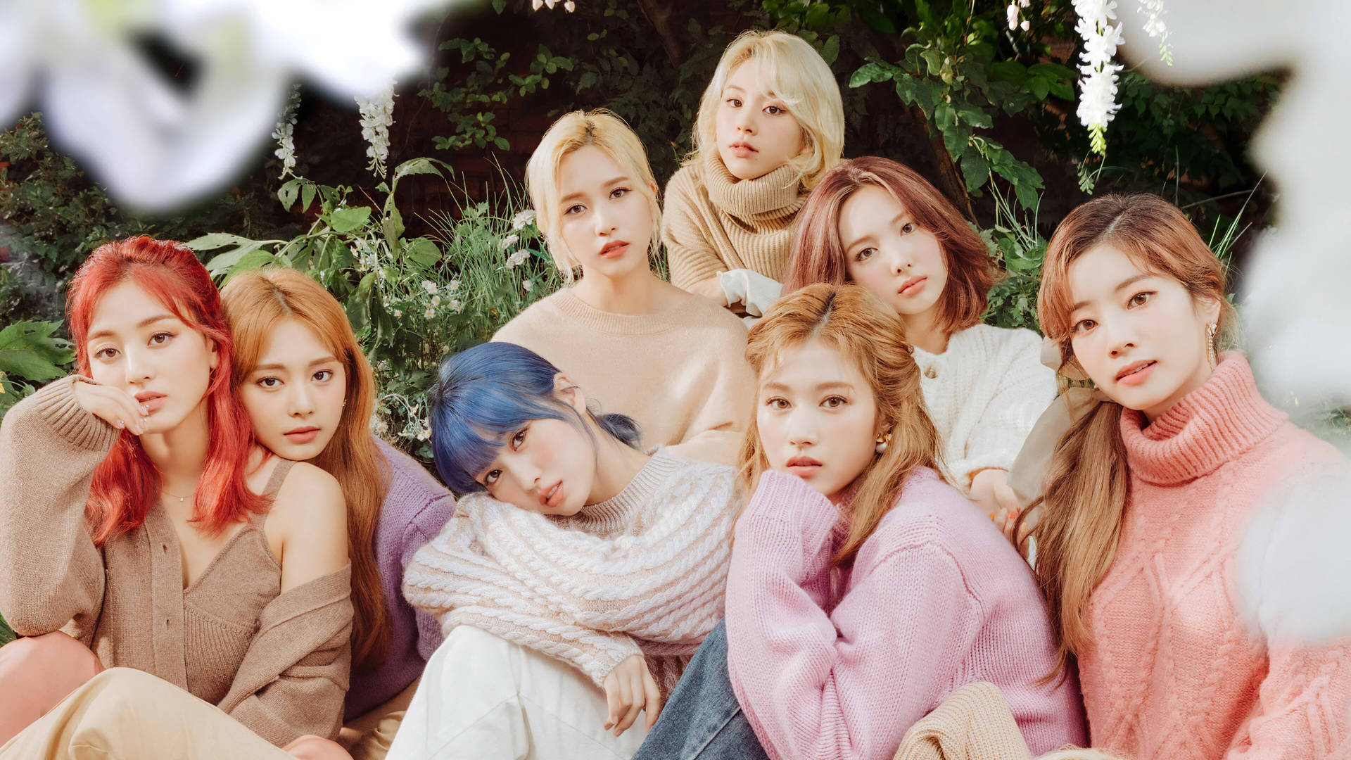 TWICE 4K 綠葉背景 壁紙 — 七位擁有繽紛髮色的女性穿著舒適毛衣，在茂密綠植與白花中一起擺姿勢。