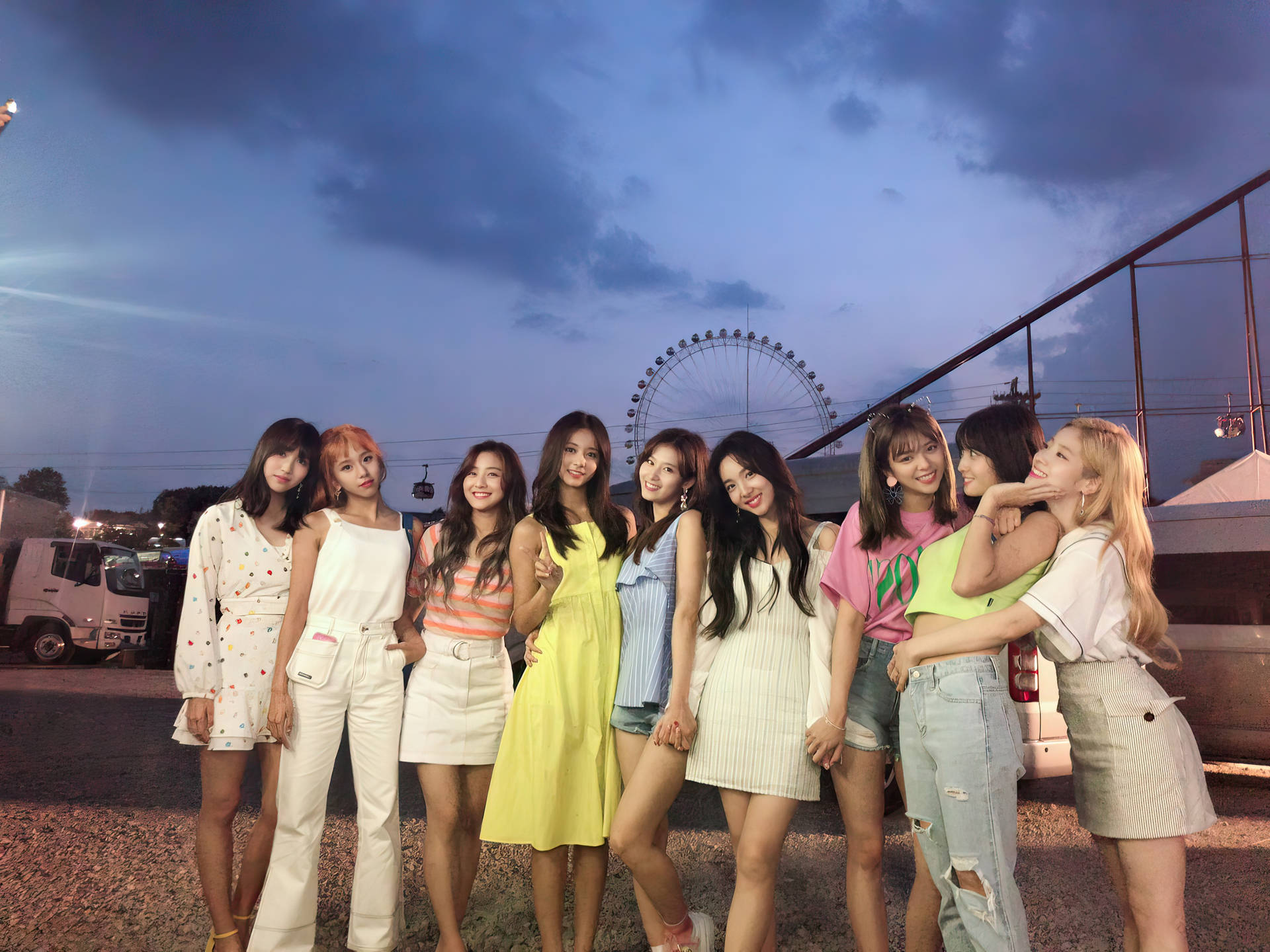 TWICE 4K 魅惑摩天輪背景 壁紙 — 九位身穿色彩繽紛夏日服裝的年輕女性，在黃昏時分於摩天輪附近，在多雲的天空下愉快地擺姿勢。