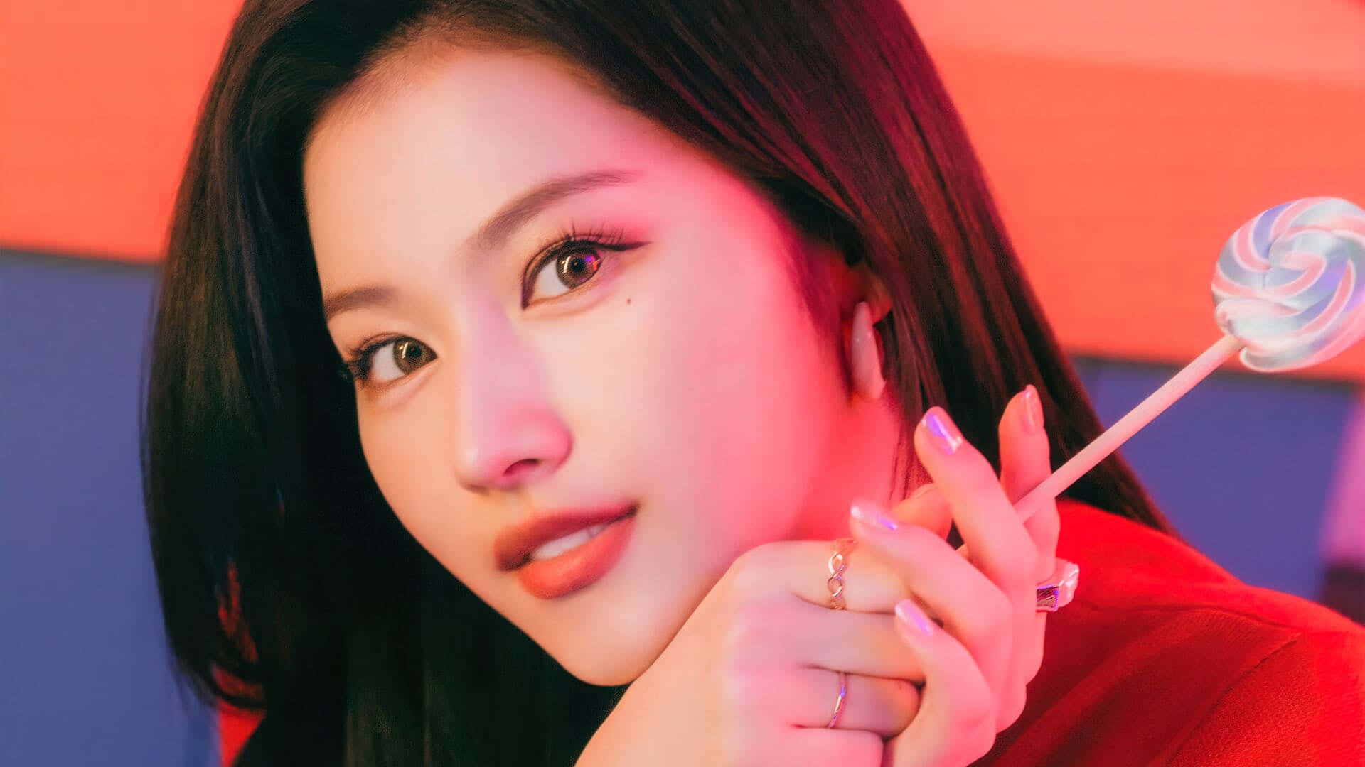 Twice K-pop 熱門天團閃耀攝影棚 壁紙 — 一位深色頭髮的年輕女性在霓虹燈光下，手持色彩繽紛的棒棒糖，背景是鮮豔的紅色和藍色。