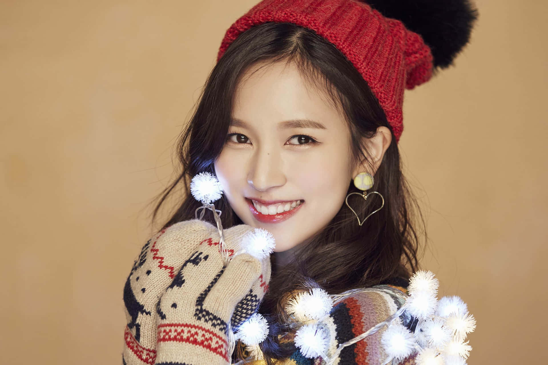 Twice K-Pop女子團體高畫質壁紙 壁紙 — 一位戴著紅色針織冷帽和圖案手套的開朗年輕女子，微笑著拿著一串白色絨球燈。