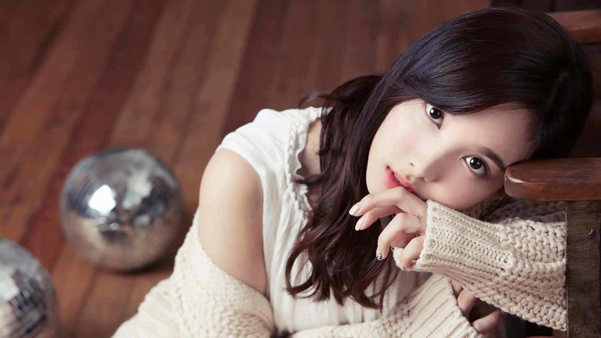 Twice專業攝影棚時尚合照 壁紙 — 一位深色頭髮的年輕女子，身穿奶油色針織毛衣，在木地板上托腮，附近有迪斯科球。