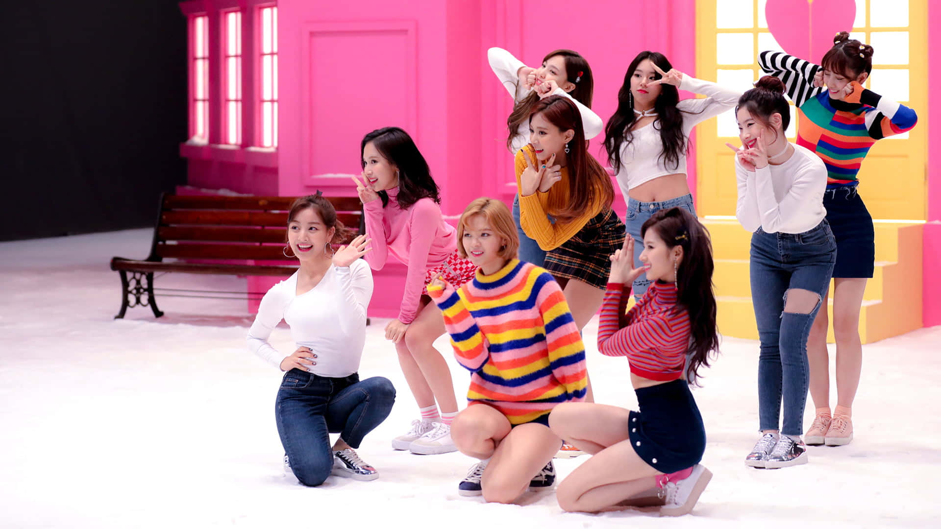 Twice 九人同台震撼演出 壁紙 — 七位身穿色彩繽紛服裝的年輕女性，在一個設有木凳的亮粉紅色和黃色場景中俏皮地擺姿勢。