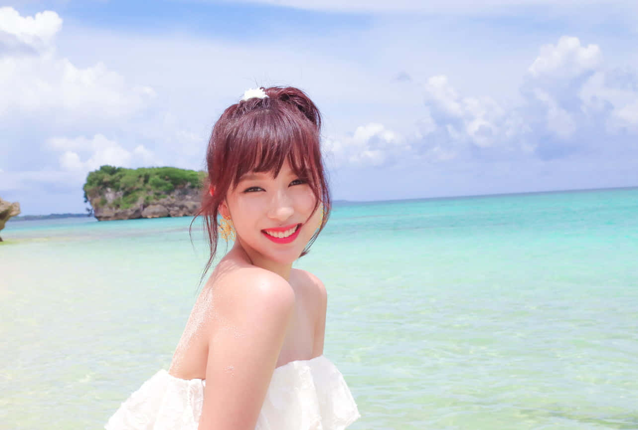 Twice - 充滿活力與時尚感的K-pop天團 壁紙 — 一位身穿白色連衣裙的快樂女士在陽光明媚的海灘上對著鏡頭微笑，背景是碧綠的海水和一個岩石島嶼。