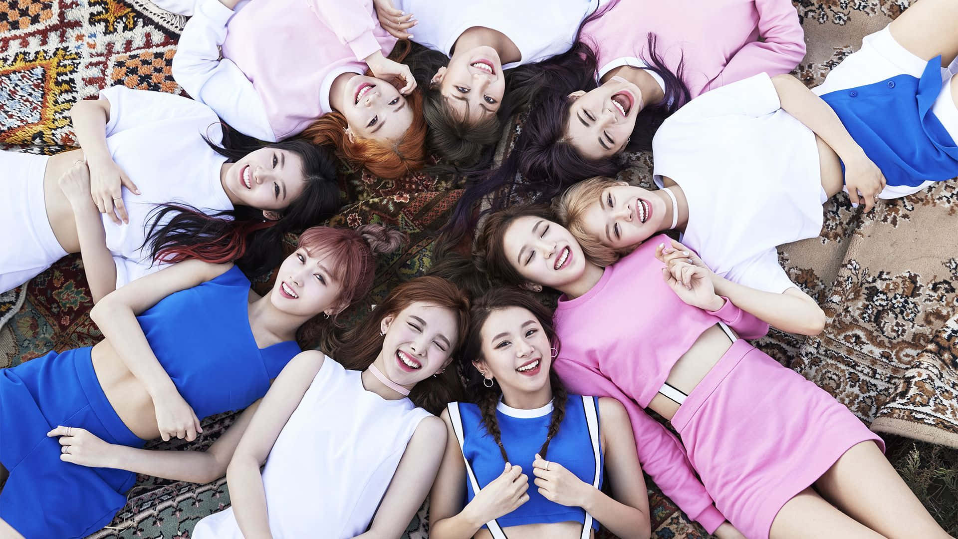 TWICE 即將以音樂震撼世界 壁紙 — 九位微笑的年輕女性身穿粉紅色和藍色服裝，躺在圖案地毯上圍成一個圓圈，開心地向上看。