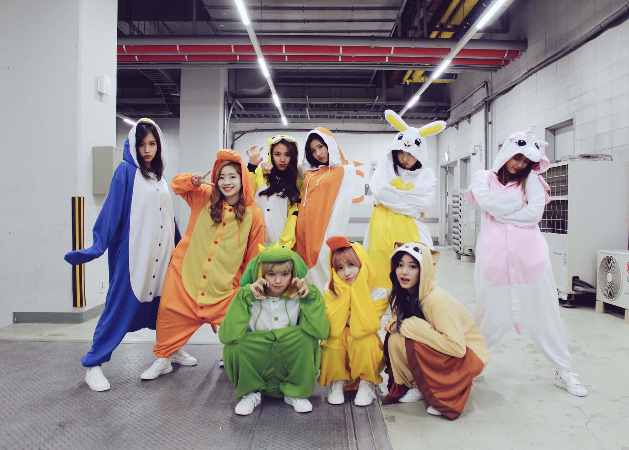 Twice 成員休閒合照 壁紙 — 七位年輕女性身穿色彩繽紛的動物連體衣，在明亮的工業車庫中俏皮地擺姿勢。