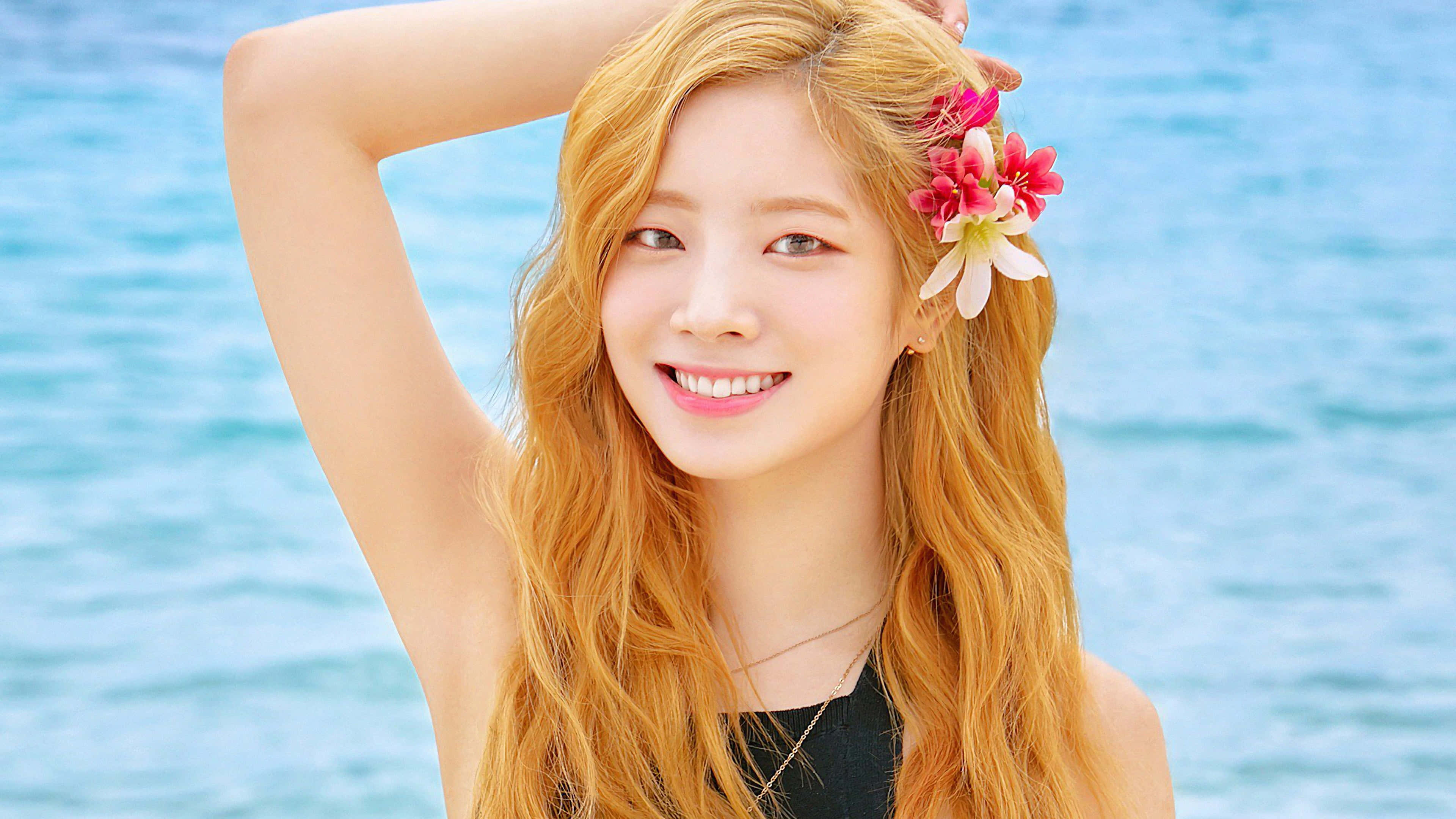 TWICE - 絕美凝視，璀璨夜色 壁紙 — 一位微笑的年輕女性，長長的金髮上點綴著粉紅色和白色的花朵，站在明亮的藍色海洋背景下。