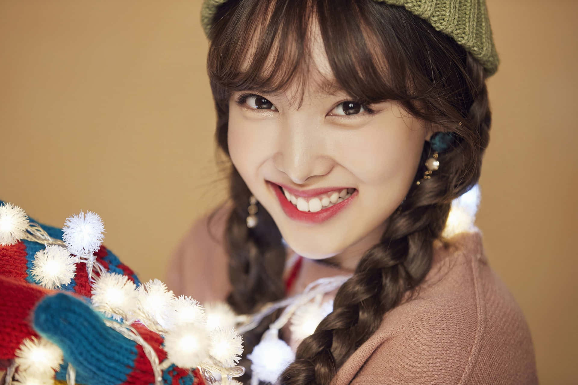 Twice 美麗攝影棚合照 壁紙 — 一位梳著辮子、戴著綠色無邊帽的開朗年輕女性，微笑著抱著一條裝飾有彩色絨球的毯子。