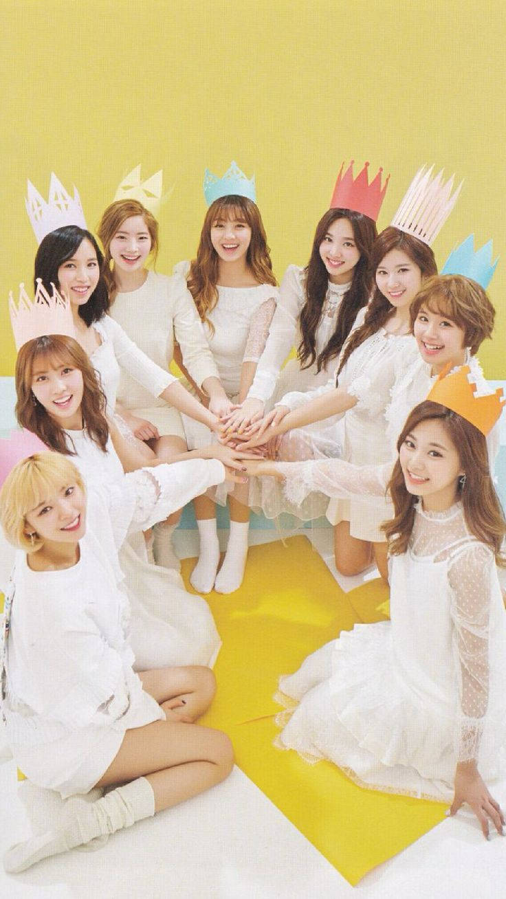 TWICE佩戴紙皇冠，甜美合影 壁紙 — 九位微笑的女性穿著白色連衣裙和彩色紙冠，在明亮的黃色背景前圍成一個圓圈疊手。