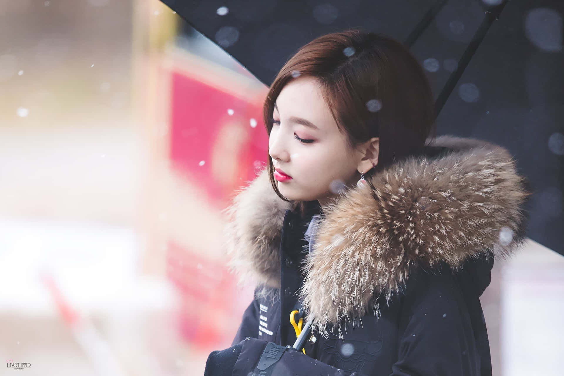 TWICE團體攝影寫真 壁紙 — 一位年輕女子穿著毛領外套，在雪花飄落的柔和模糊城市背景下，撐著一把黑色雨傘。