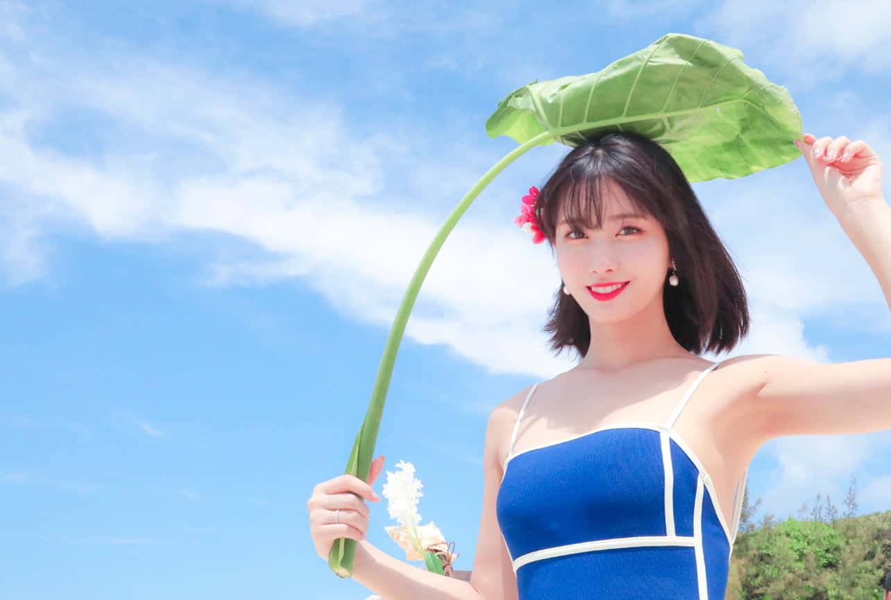 TWICE成員拍攝迷人團體照 壁紙 — 一位身穿藍色連衣裙的微笑女子，在明亮的藍天和蓬鬆雲朵下，將一片巨大的綠葉舉過頭頂。