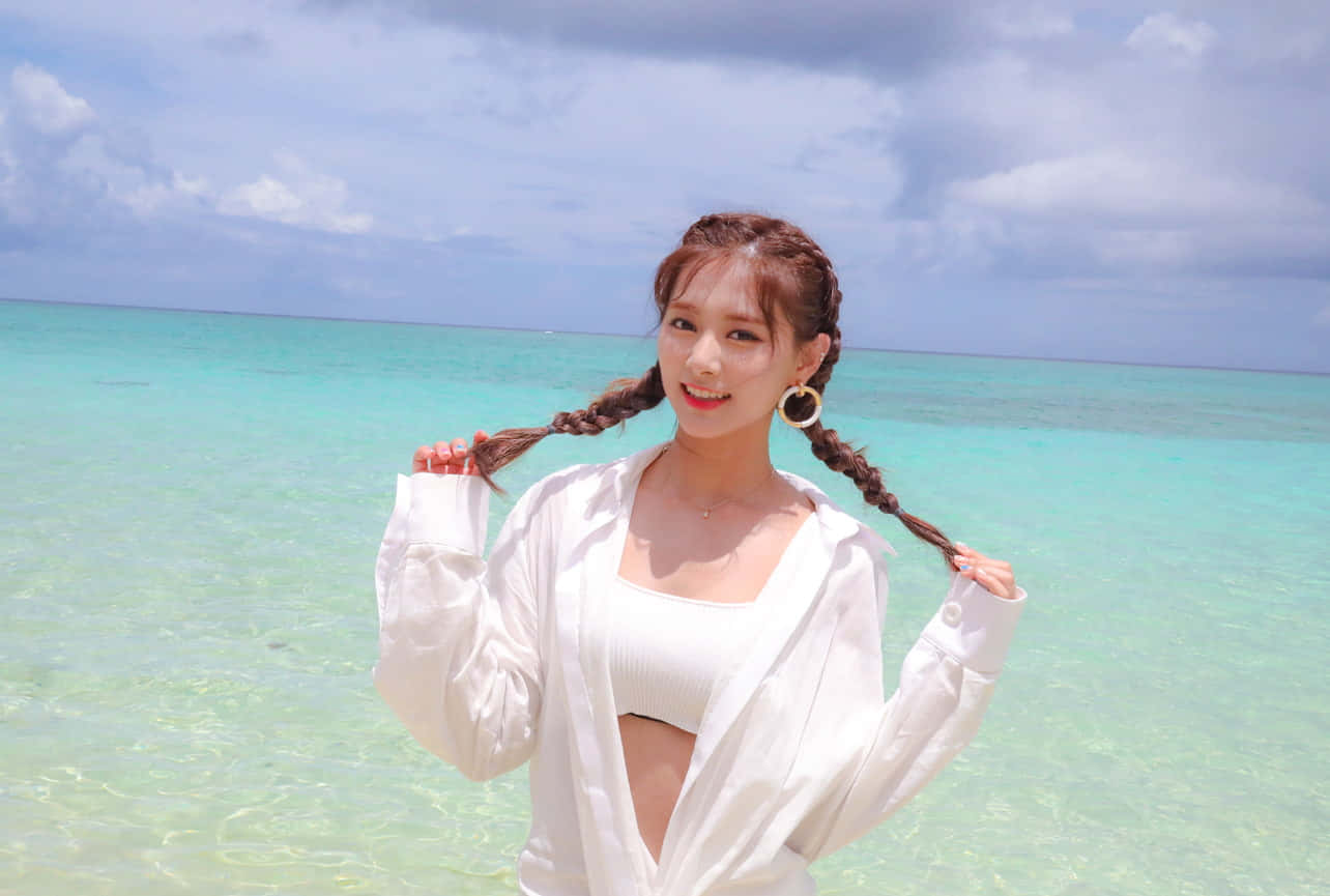 TWICE成員身穿優雅服裝擺出可愛姿勢 壁紙 — 一位微笑的辮子女性在部分多雲的天空下，在碧綠海洋的邊緣頑皮地握著她的辮子。