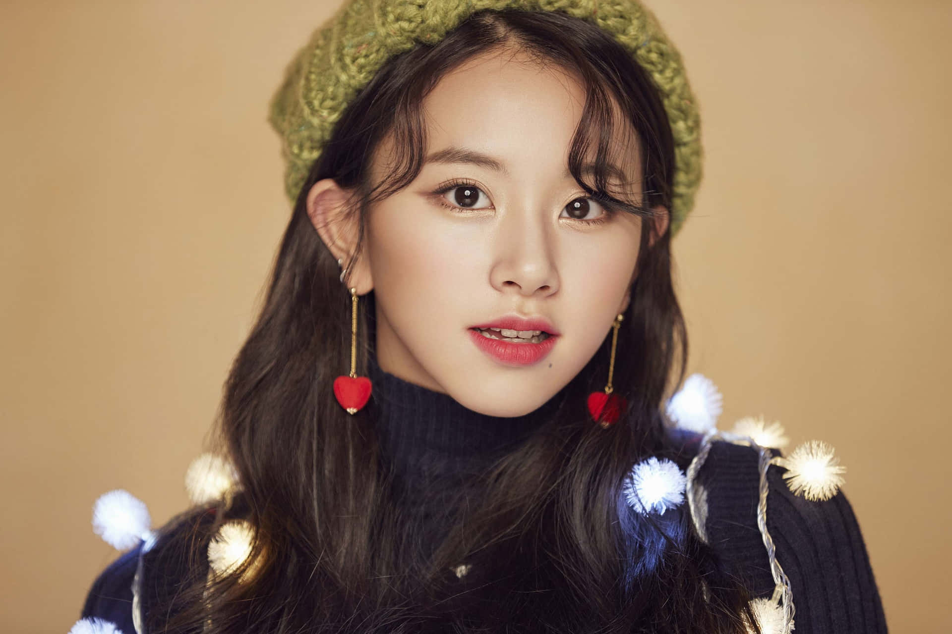 TWICE穿時尚Vogue服裝團體照 壁紙 — 一位長黑髮的年輕女性，在溫暖的米色背景前，戴著綠色針織貝雷帽，穿著飾有白色絨球和紅色心形耳環的深色毛衣。