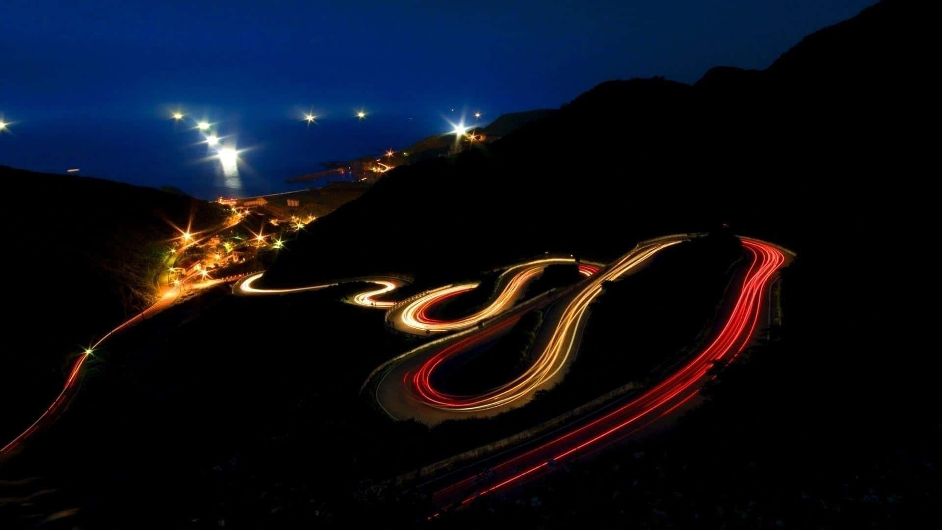  壁紙 — 夜晚蜿蜒的海岸道路，被沿著黑暗山坡蜿蜒行駛的汽車發出的發光光跡照亮。