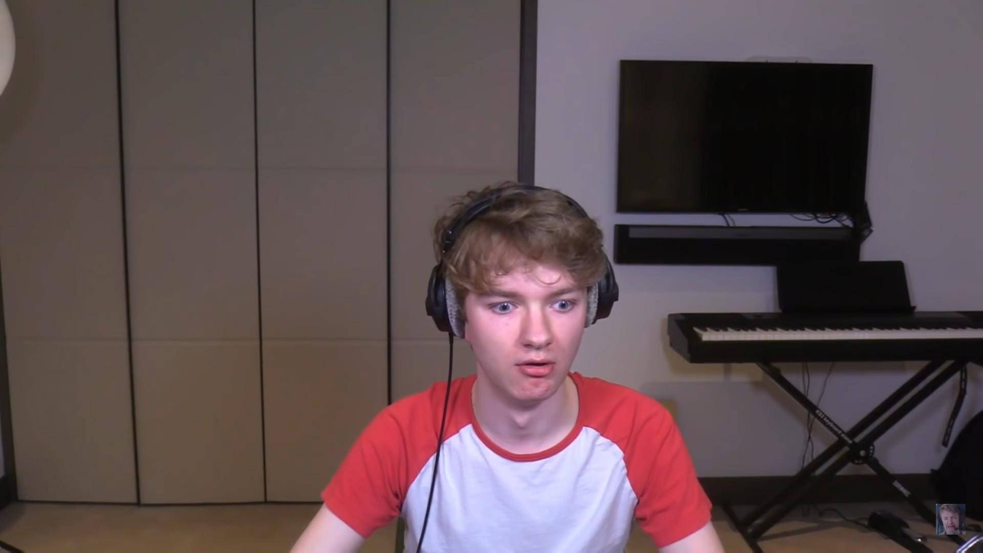 Twitch直播主Tommyinnit 壁紙 — 年輕男子戴著耳機，在家中的錄音室裡，身後有鍵盤和電視，驚訝地盯著鏡頭。