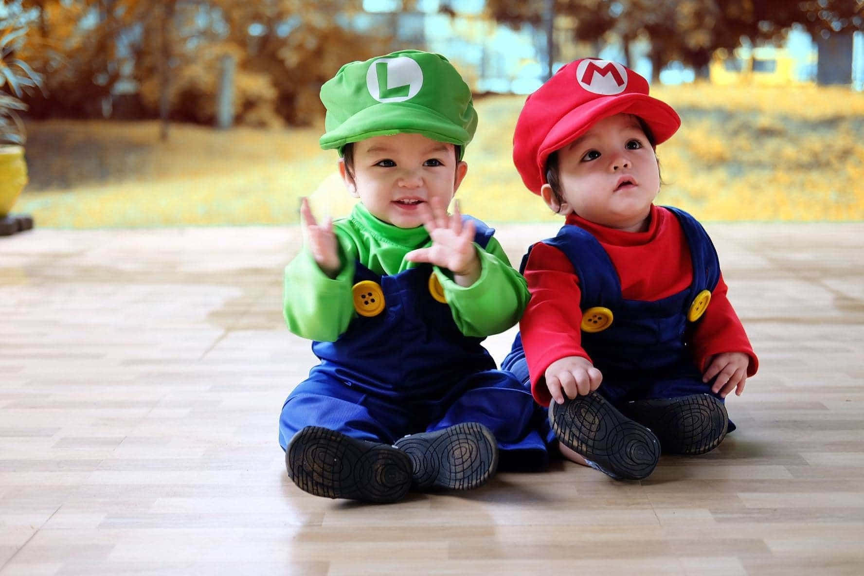 Baby Luigi 桌布 壁紙 — 兩個可愛嘅BB扮成孖寶兄弟，坐喺木地板上面開心咁向鏡頭揮手。