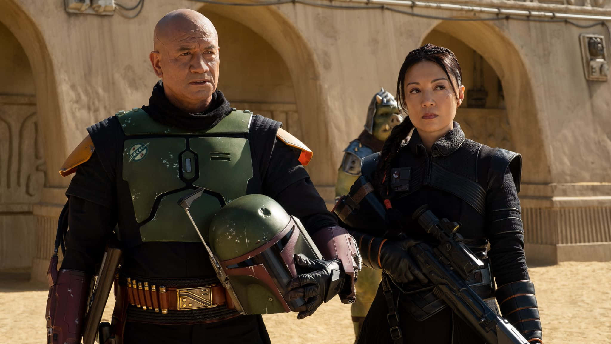 Ming Na Wen 桌布 壁紙 — 兩名身穿盔甲的賞金獵人在陽光明媚的沙漠小鎮中保持警覺，身後是拱形石頭建築。