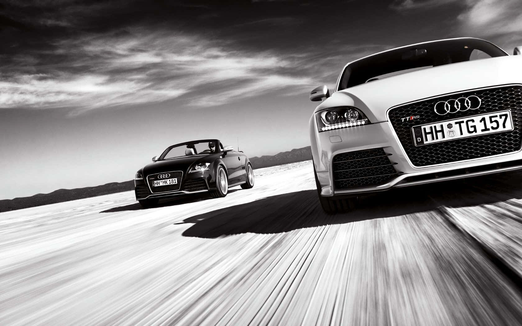 Audi Tt Rs 桌布 壁紙 — 兩輛奧迪跑車在廣闊的沙漠中競速，動態模糊強調了速度，背景是戲劇性的天空。