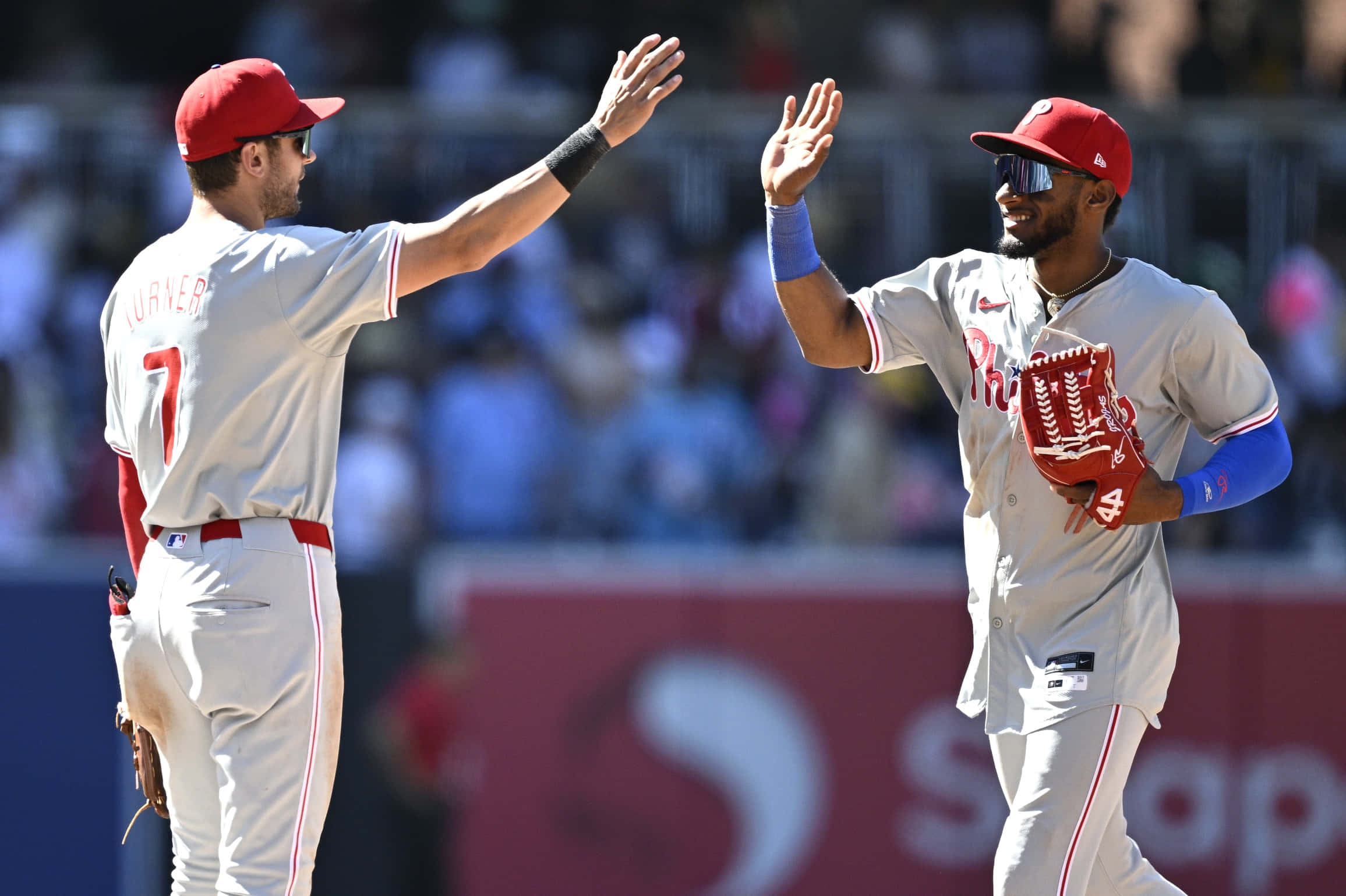 Trea Turner Phillies 桌布 壁紙 — 兩名身穿灰色和紅色球衣的棒球運動員在陽光明媚的球場上擊掌，背景是模糊的人群。