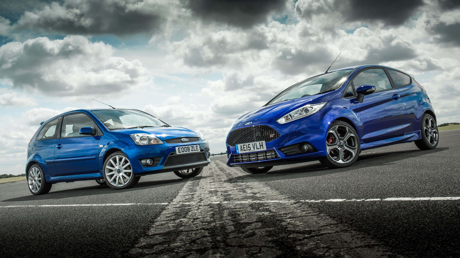 Ford Fiesta 桌布 壁紙 — 兩輛藍色福特Fiesta掀背車在戲劇性的多雲天空下，在潮濕的瀝青路上對峙。