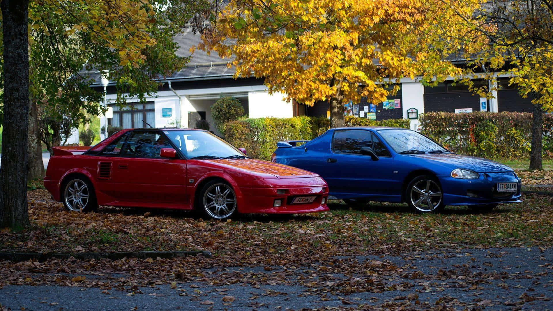 Honda Civic Del Sol 桌布 壁紙 — 兩輛經典跑車，紅色豐田MR2和藍色本田S2000，停泊在金色秋樹下，落葉覆蓋地面。