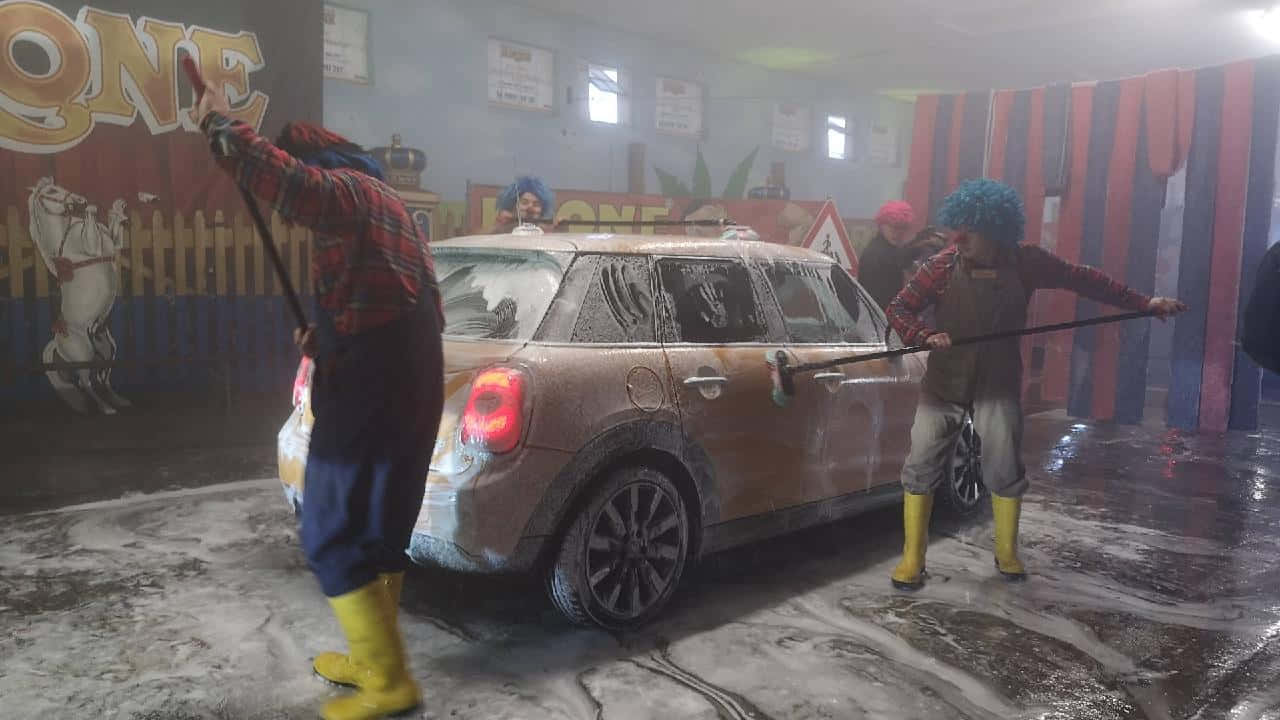  壁紙 — 兩個穿著亮黃色靴子的小丑在色彩繽紛的室內洗車場擦洗一輛滿是肥皂泡的汽車，洗車場內有條紋窗簾和壁畫。