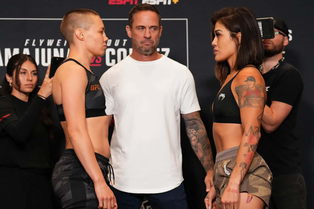  壁紙 — 兩位女性MMA拳手在明亮的攝影棚燈光下，於稱重儀式上對峙，中間站著一位裁判。