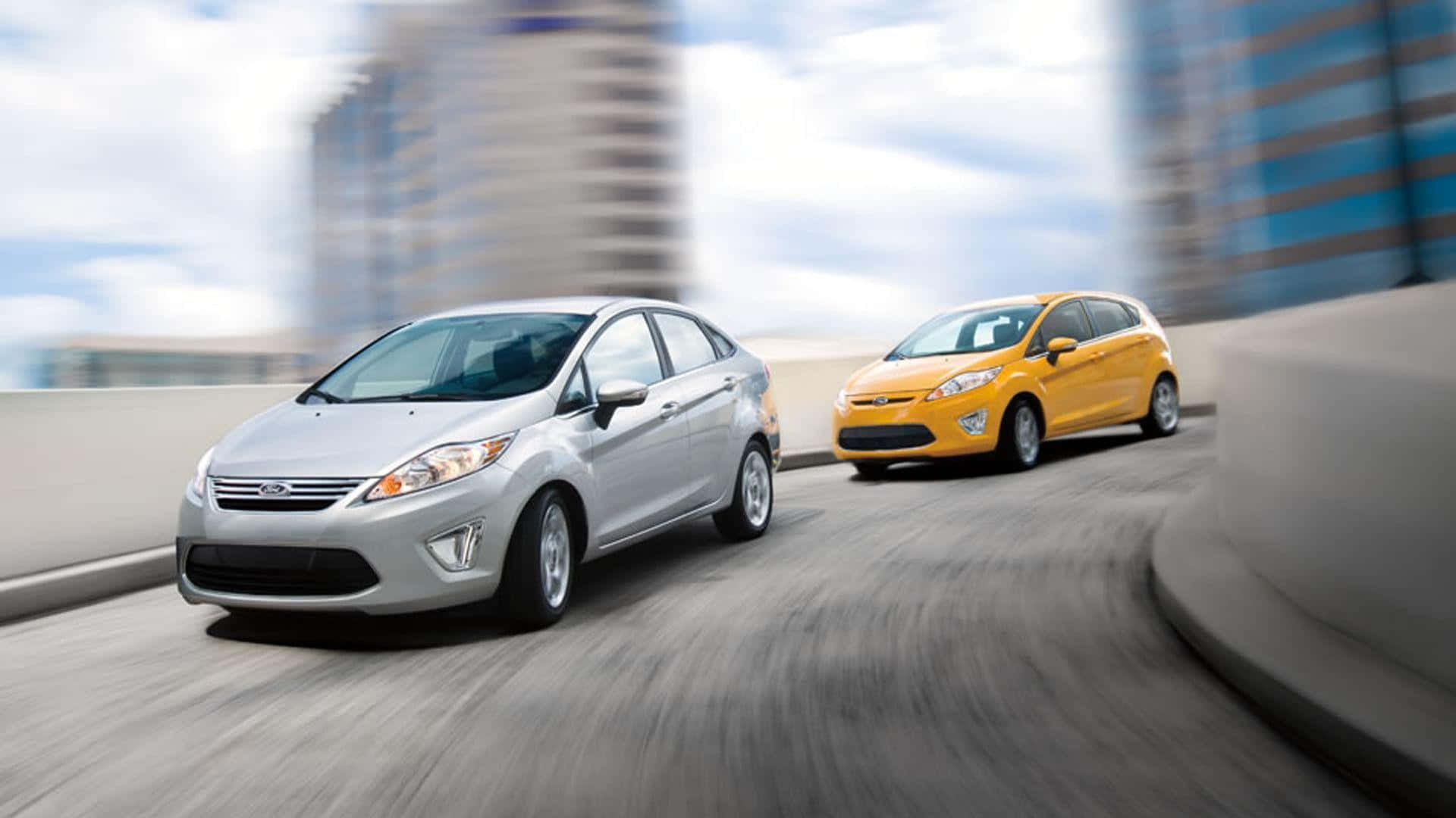Ford Fiesta 桌布 壁紙 — 兩輛福特汽車，銀色和黃色，沿著彎曲的城市高架橋奔馳，帶有動態模糊效果，背景是城市天際線。