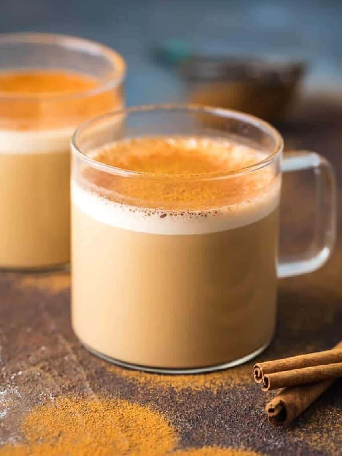  壁紙 — 兩杯玻璃杯裝的奶油香料拿鐵，上面撒有肉桂粉和整根肉桂棒，旁邊是質樸的木質表面。