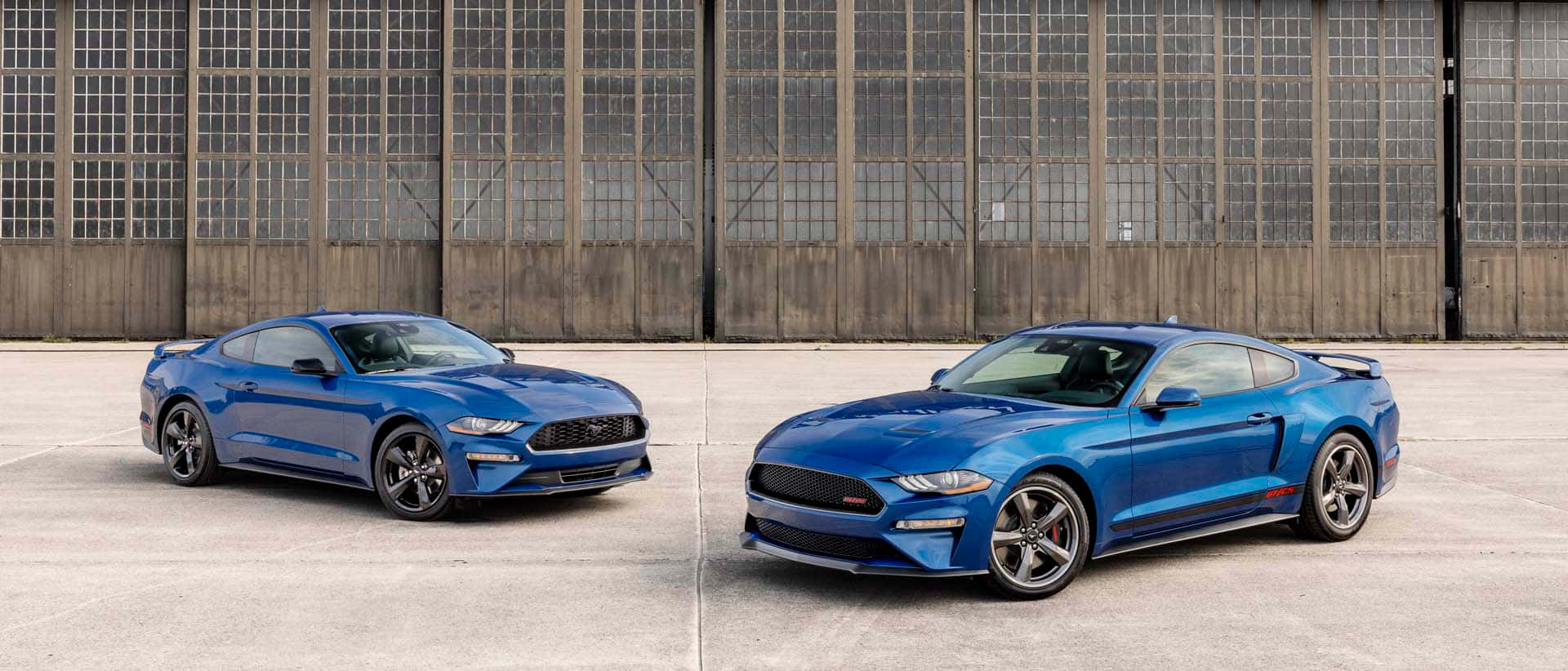 Ford Mustang California Special 桌布 壁紙 — 兩輛閃亮的藍色福特野馬在一個帶有格柵窗戶的大型工業機庫前相對而立。