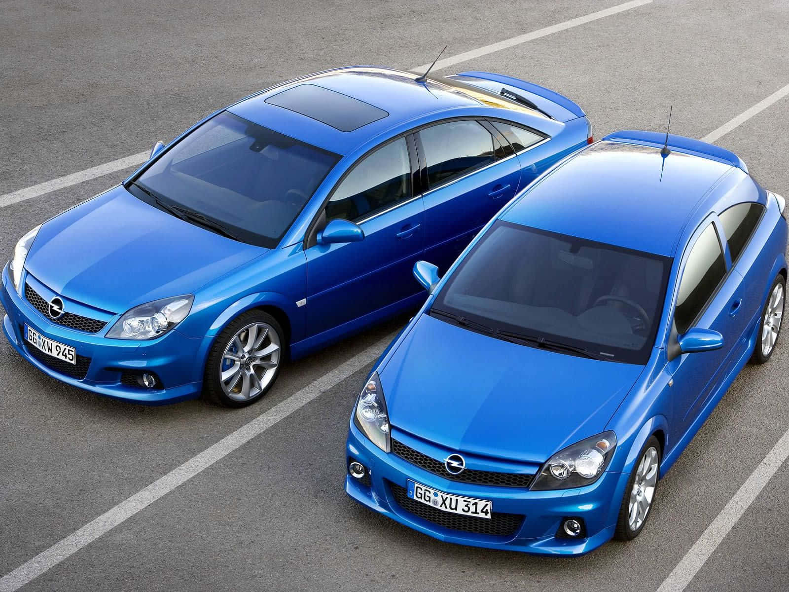 Opel Astra 桌布 壁紙 — 兩輛亮藍色歐寶跑車在明亮的日光下並排停放在灰色瀝青場地上。