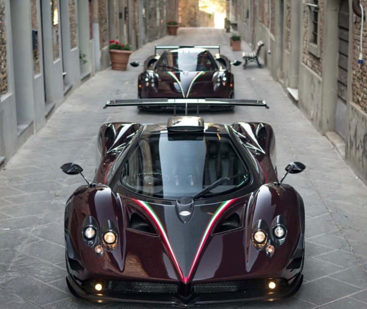 Pagani Zonda Tricolore 桌布 壁紙 — 兩輛光澤栗色Pagani超跑帶紅色點綴，停放在歷史建築之間的狹窄石巷中。