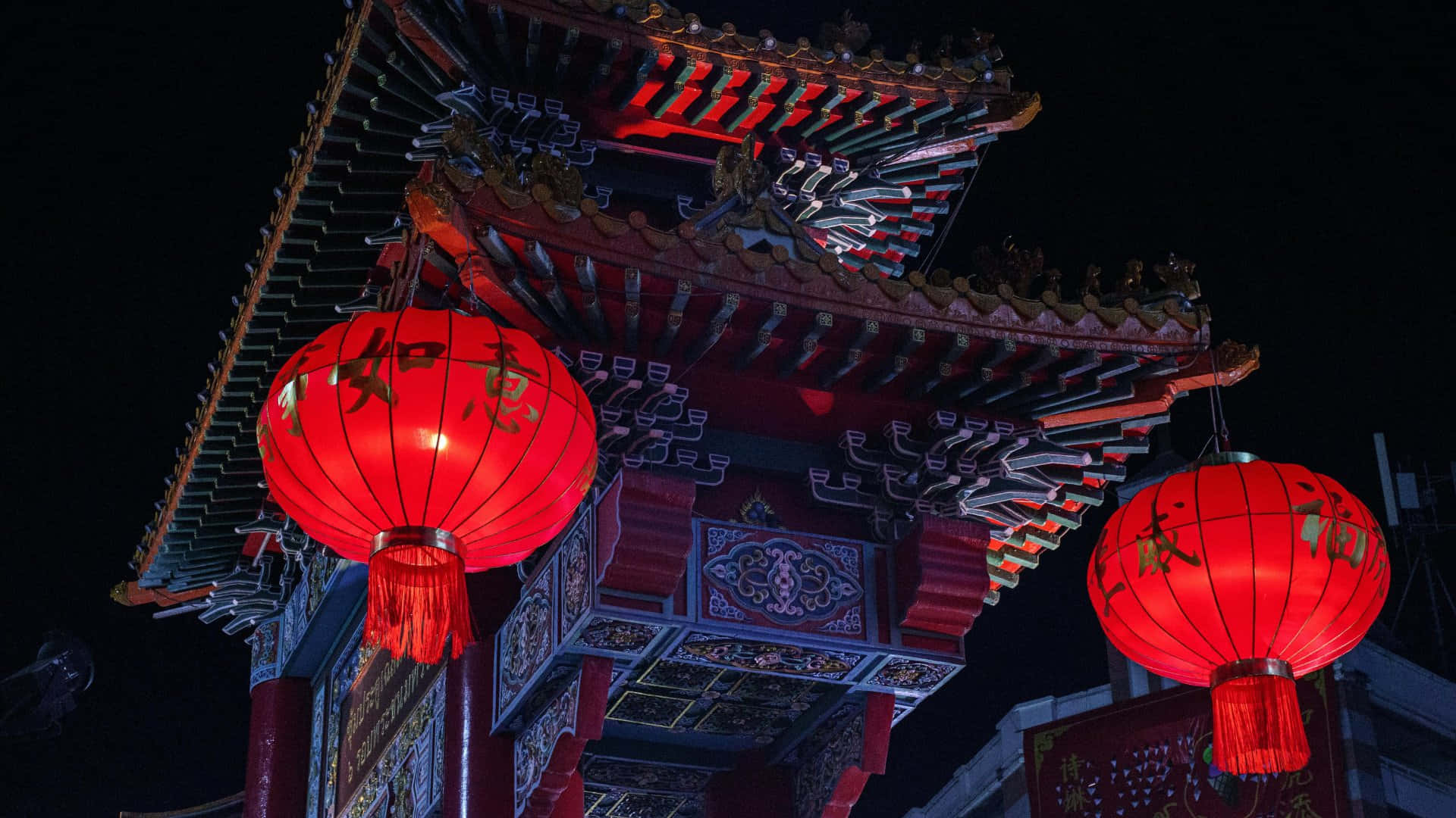  壁紙 — 兩個發光的紅色中國燈籠掛在一個華麗的傳統亭子前，背景是漆黑的夜空。