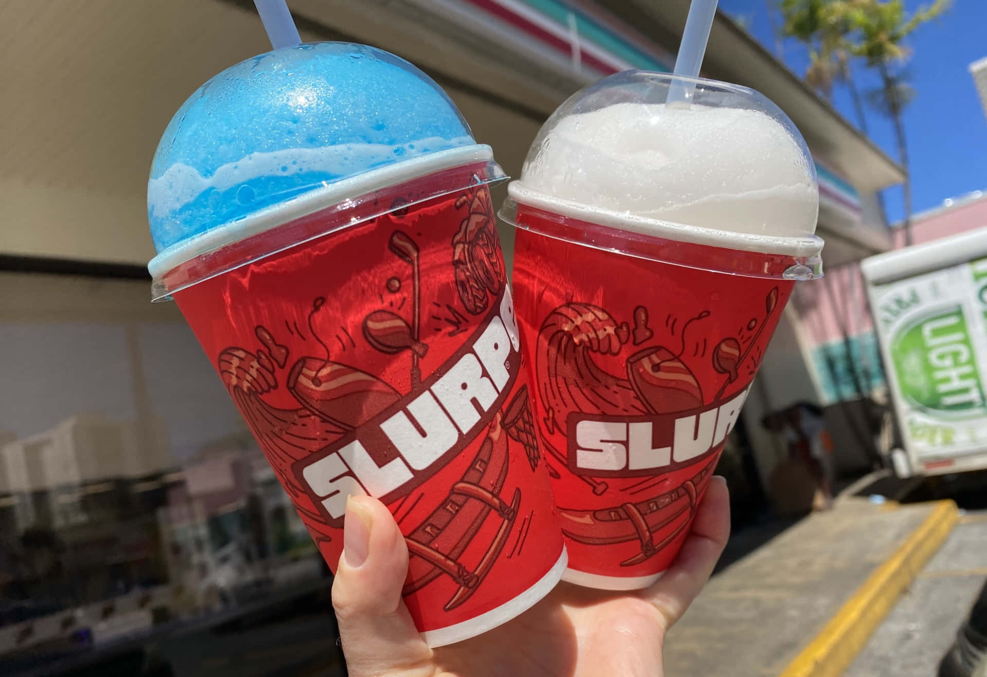  壁紙 — 兩隻手拿著鮮紅色的 Slurpee 杯，杯上有藍白相間的泡沫頂部，背景是陽光明媚的街道。