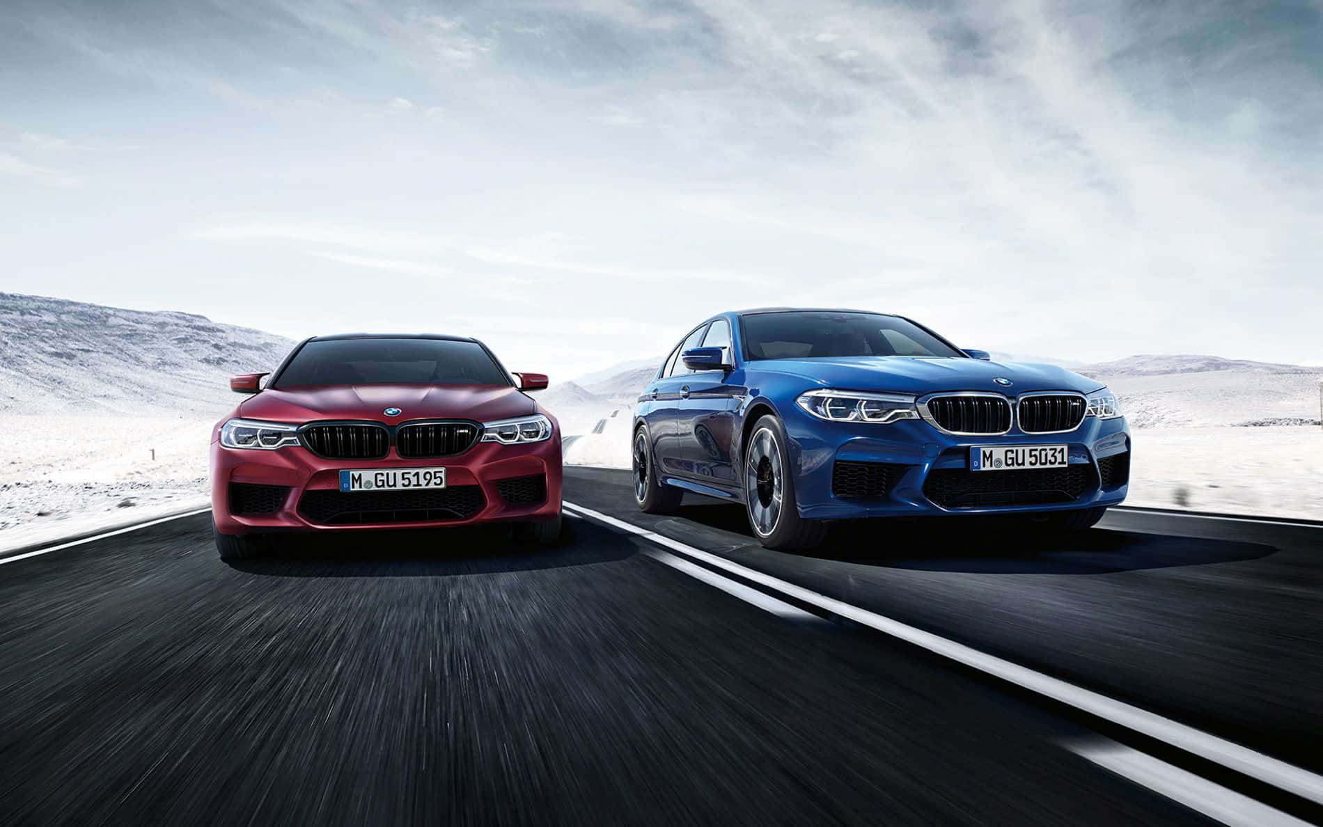 Bmw M5 桌布 壁紙 — 兩輛高性能紅色和藍色BMW M5房車在多雲的天空下，在積雪的山路上並排競速。