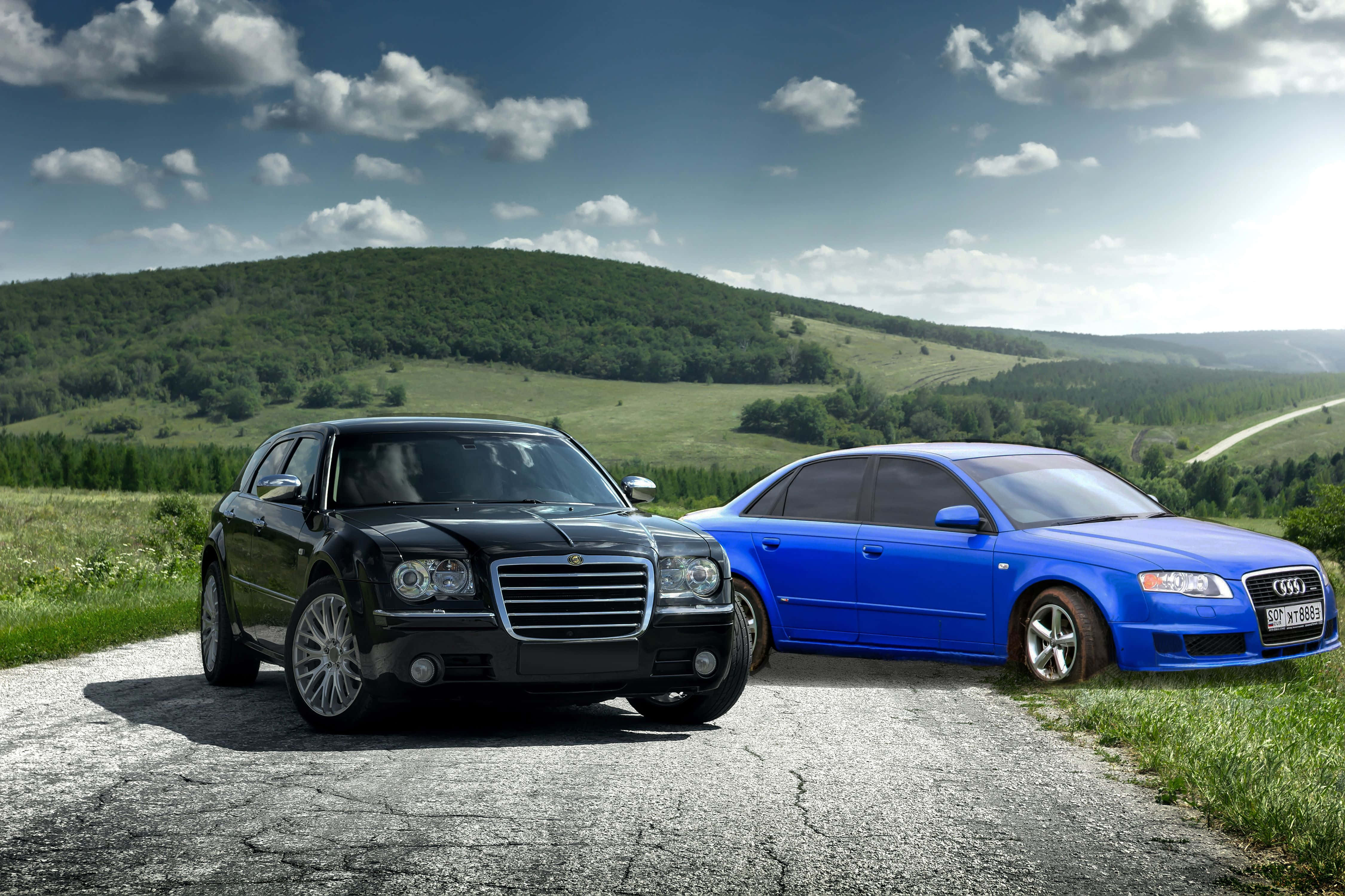 Chrysler 300 桌布 壁紙 — 兩輛豪華汽車黑色和藍色停在一條裂開的道路上，背景是連綿起伏的綠色山丘和部分多雲的天空。