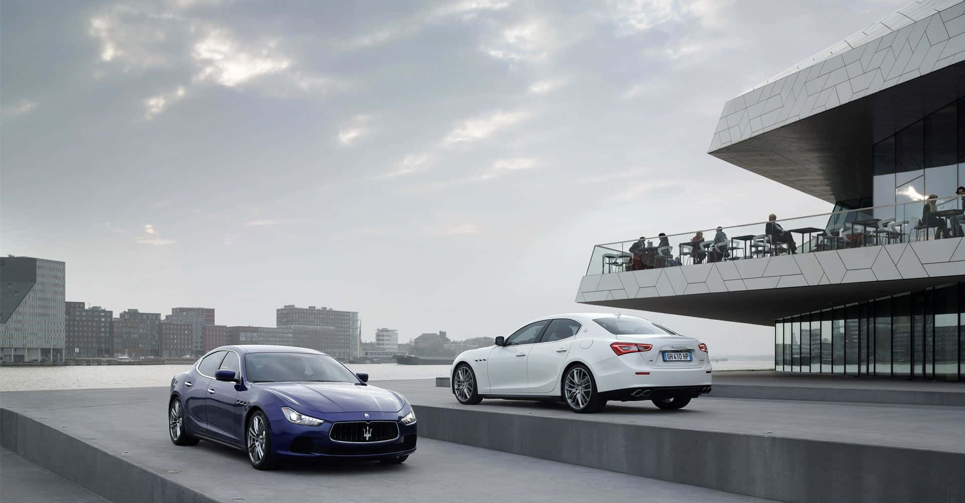 Maserati Ghibli 桌布 壁紙 — 兩輛豪華瑪莎拉蒂房車停泊在現代海濱廣場上，背景是玻璃幕牆建築和城市天際線，天空多雲。