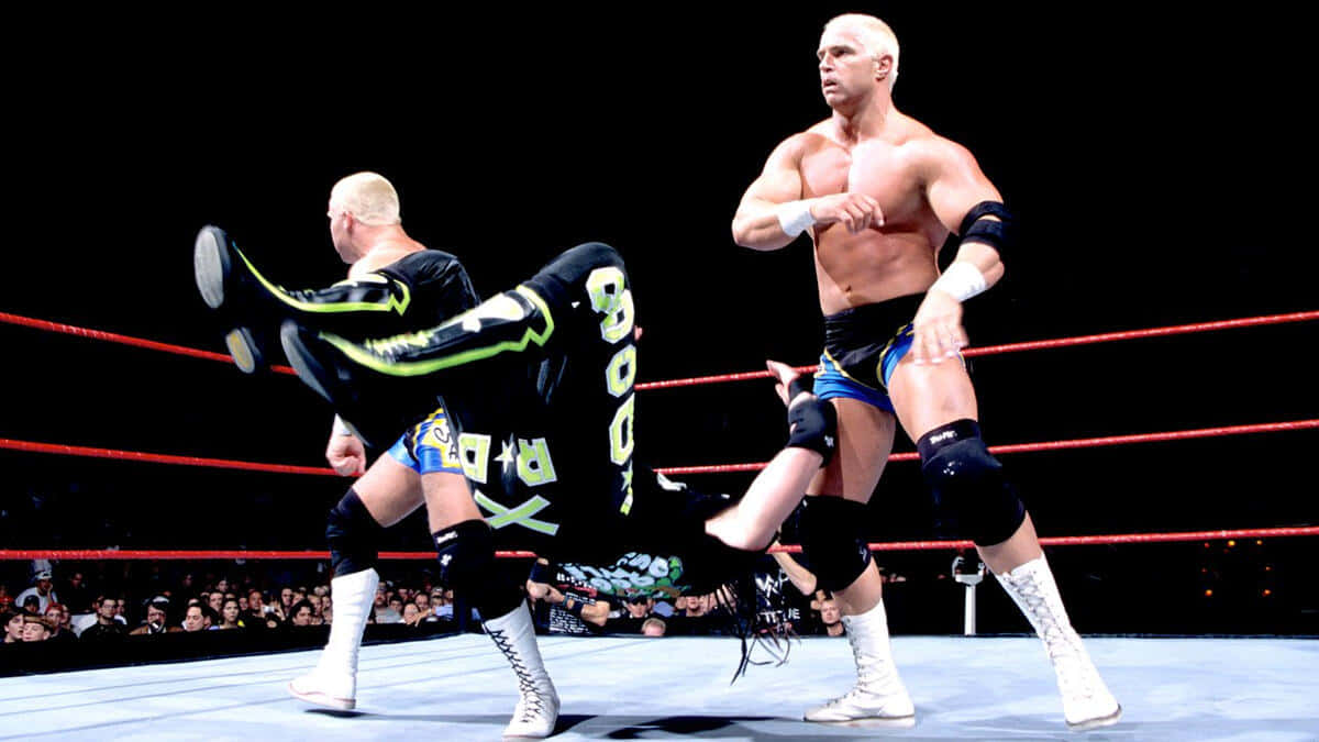 Crash Holly 桌布 壁紙 — 兩名肌肉發達的摔角手身穿黑色和藍色裝備，在燈光明亮、有紅色繩索和歡呼人群的擂台上激烈纏鬥。