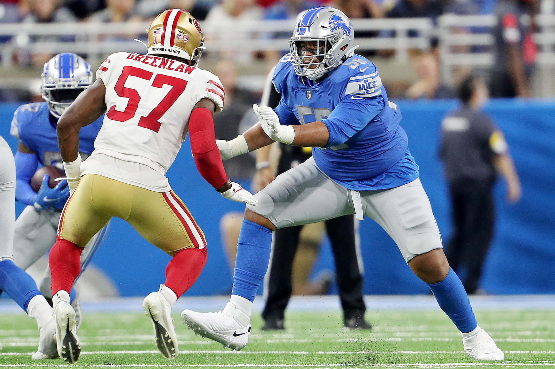  壁紙 — 兩名身穿藍色和金色球衣的NFL球員在明亮的體育場燈光下，於夜間比賽中在綠色球場上激烈碰撞。