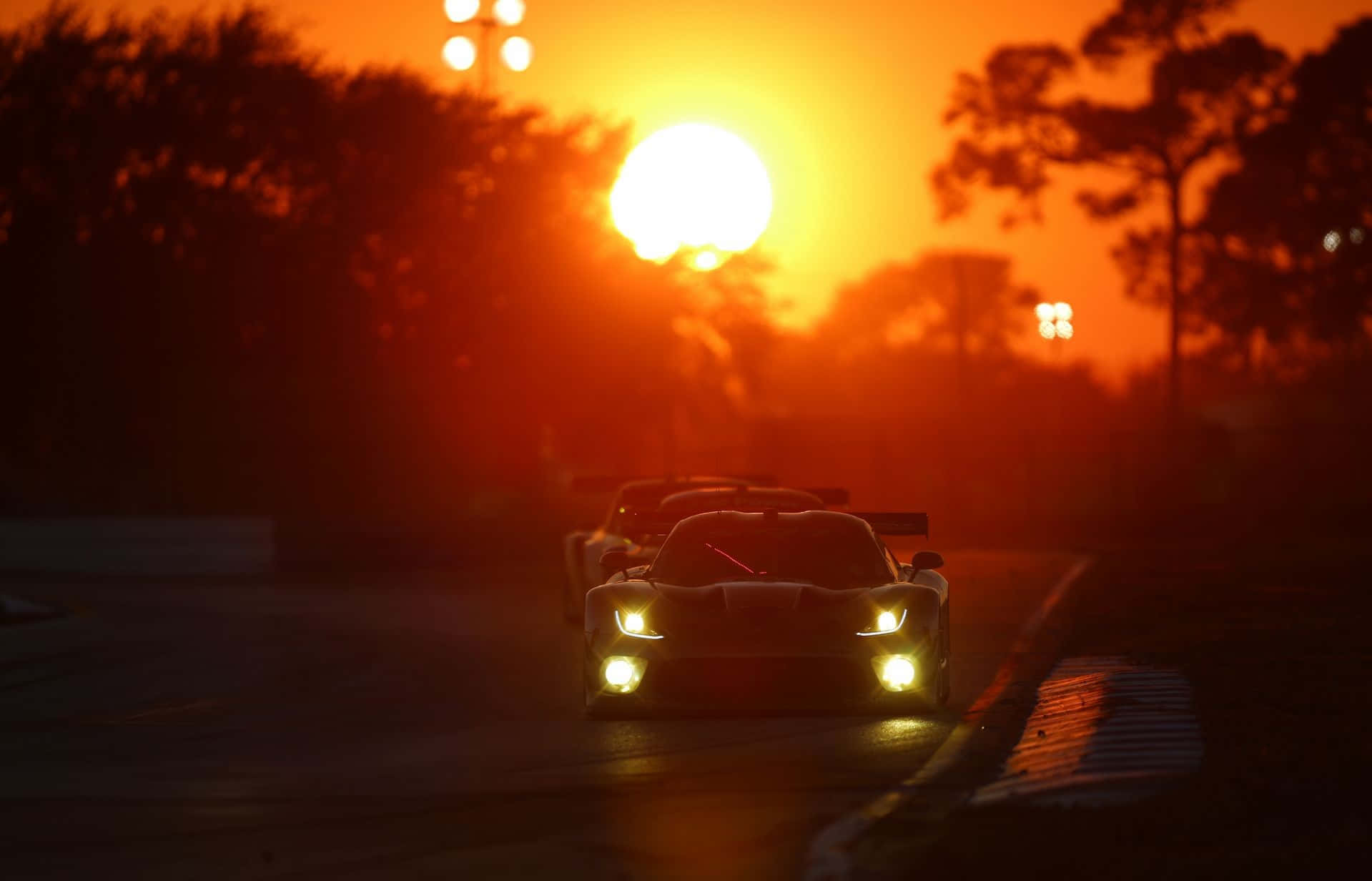 Sunset Drive 桌布 壁紙 — 兩輛賽車在日落時沿著賽道滑行，發光的車頭燈劃破熾熱的橙色光芒。