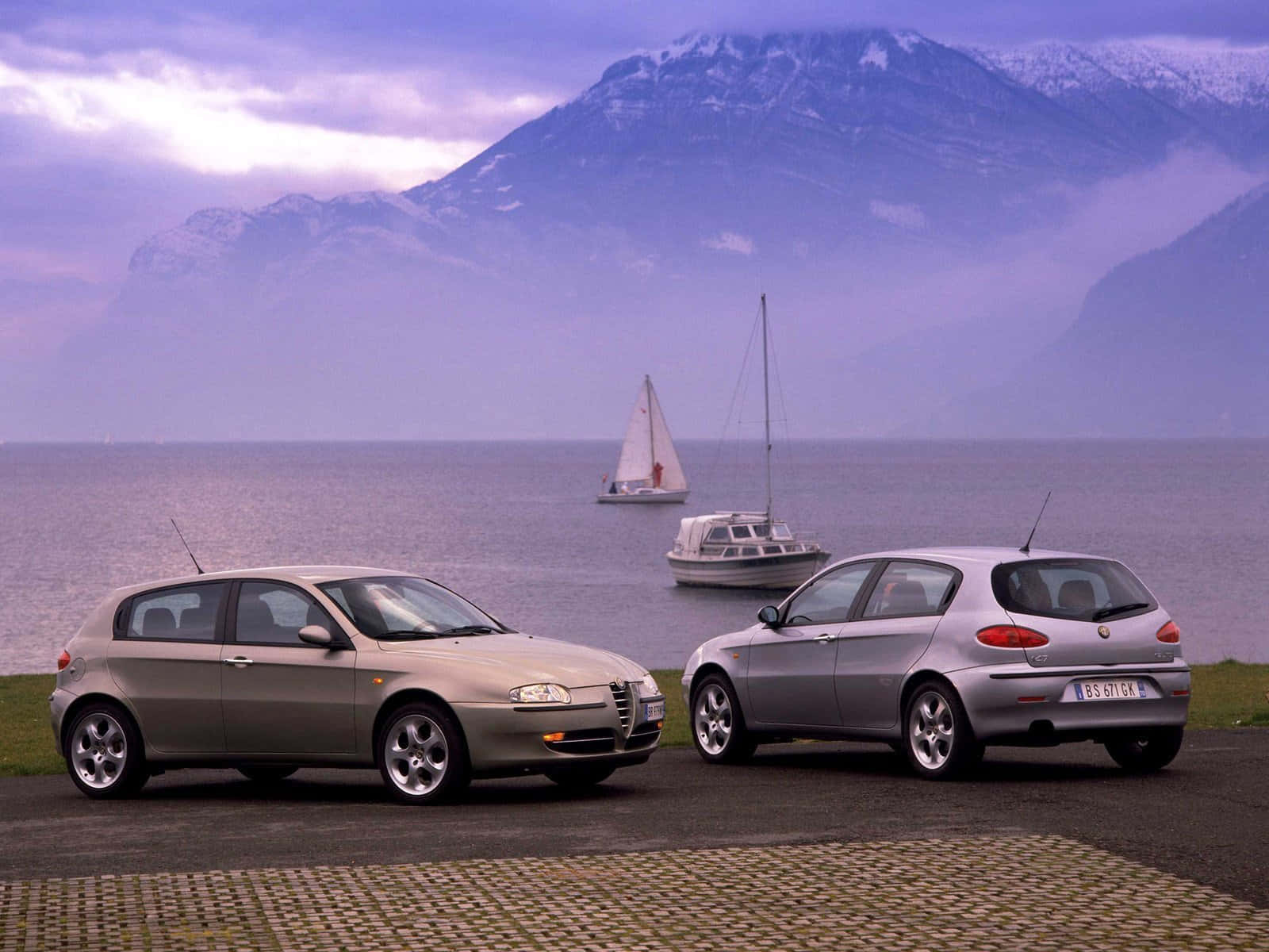 Alfa Romeo 147 桌布 壁紙 — 兩輛銀色愛快羅密歐汽車停泊在湖邊，湖上有帆船，背景是白雪覆蓋的山脈，天空呈紫色。