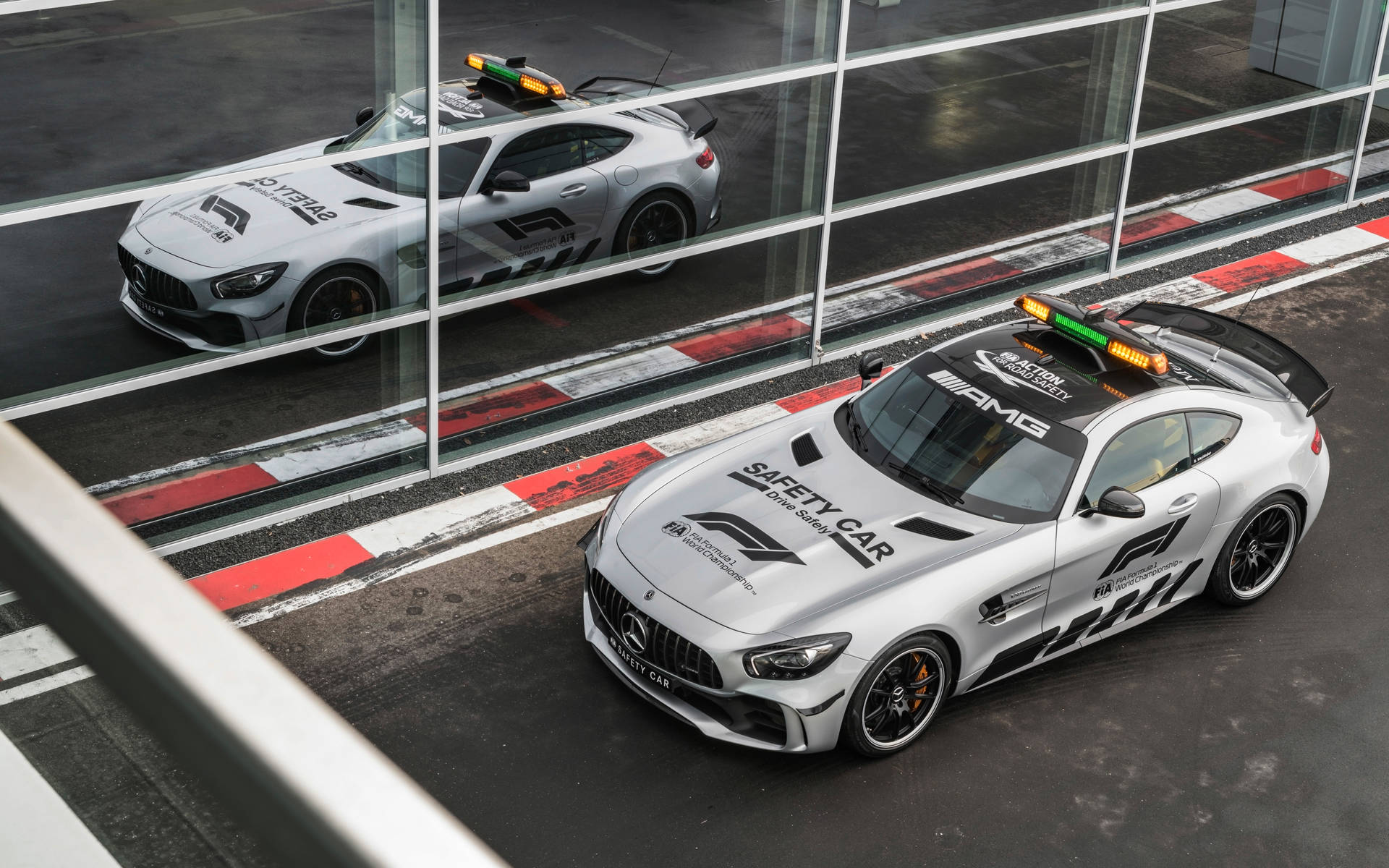 AMG GTR 賽道窗面倒影 壁紙 — 兩輛銀色梅賽德斯-AMG GT安全車，閃爍著燈光，停在賽道旁的玻璃屏障邊。